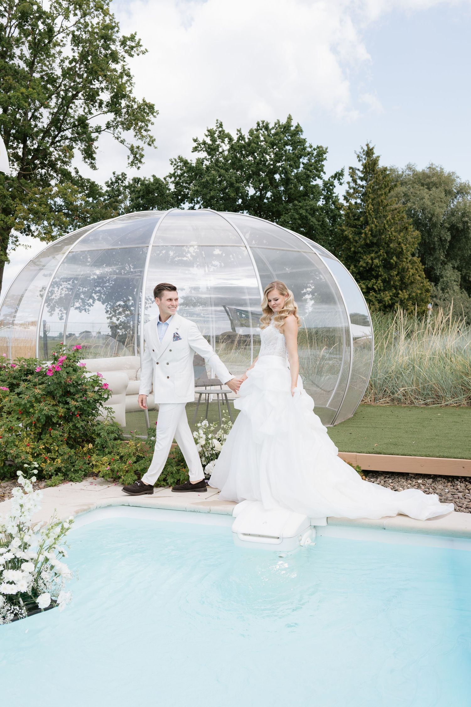 Bride and groom in white attire hold hands by a pool near a transparent dome structure outdoors.