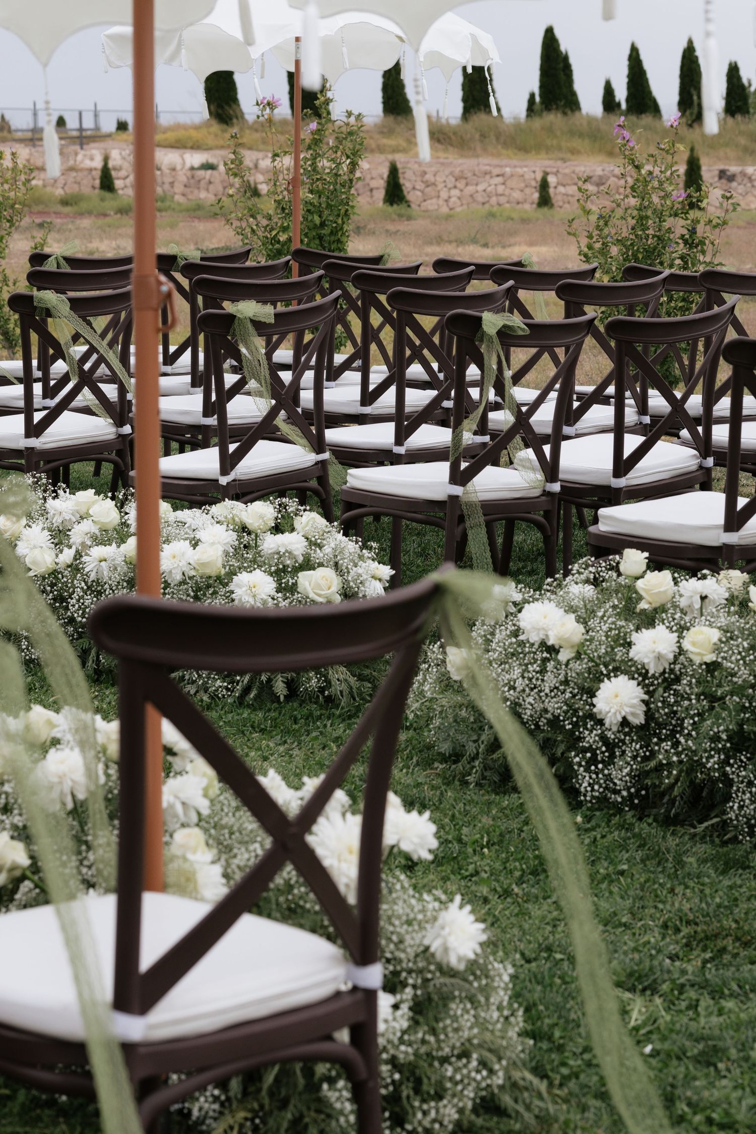 Outdoor wedding ceremony setup with dark wood cross-back chairs, white cushions, and white floral arrangements on green grass aisle.