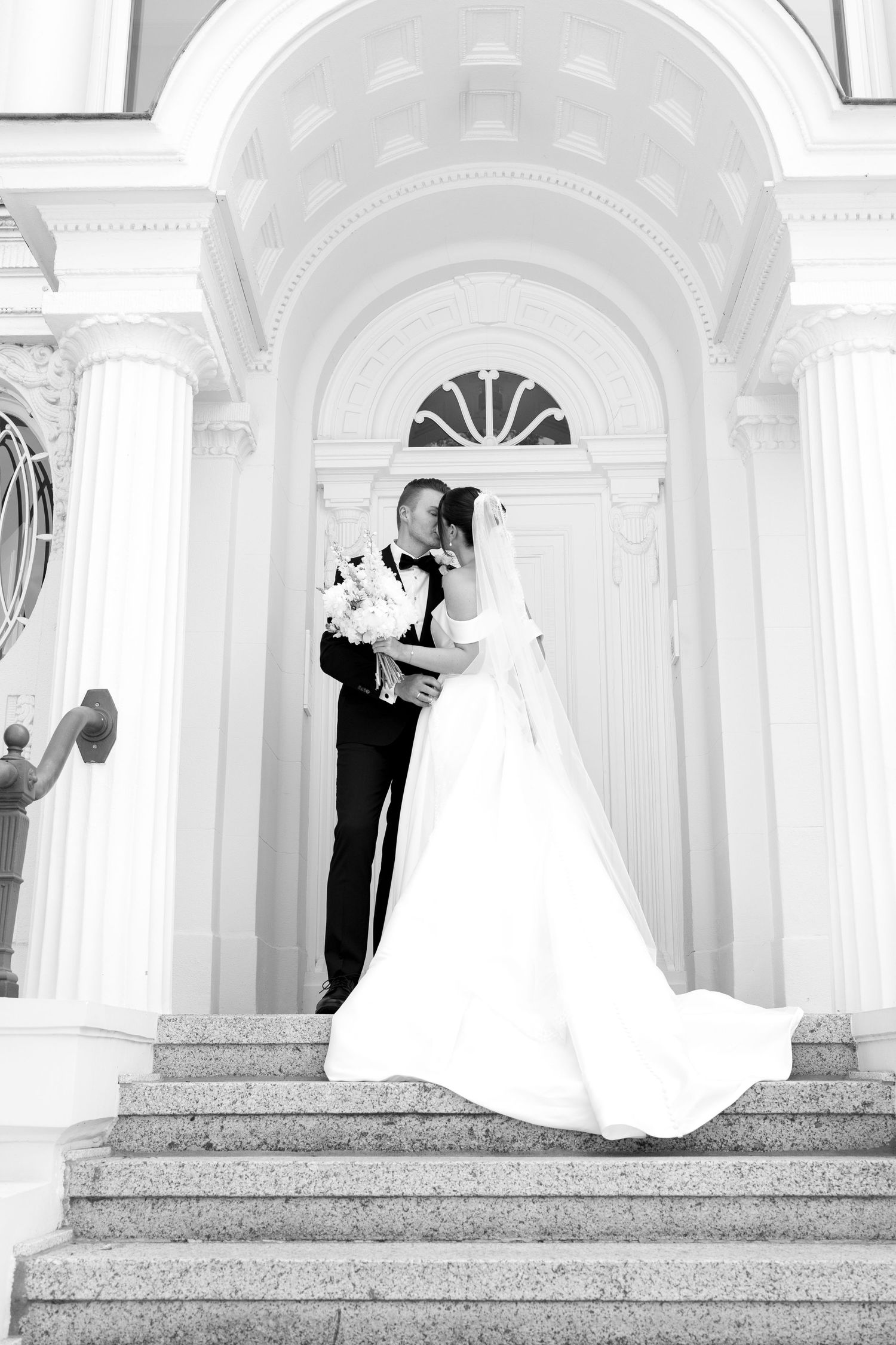 Black and white photo of bride and groom kissing on grand staircase with elegant white columns.