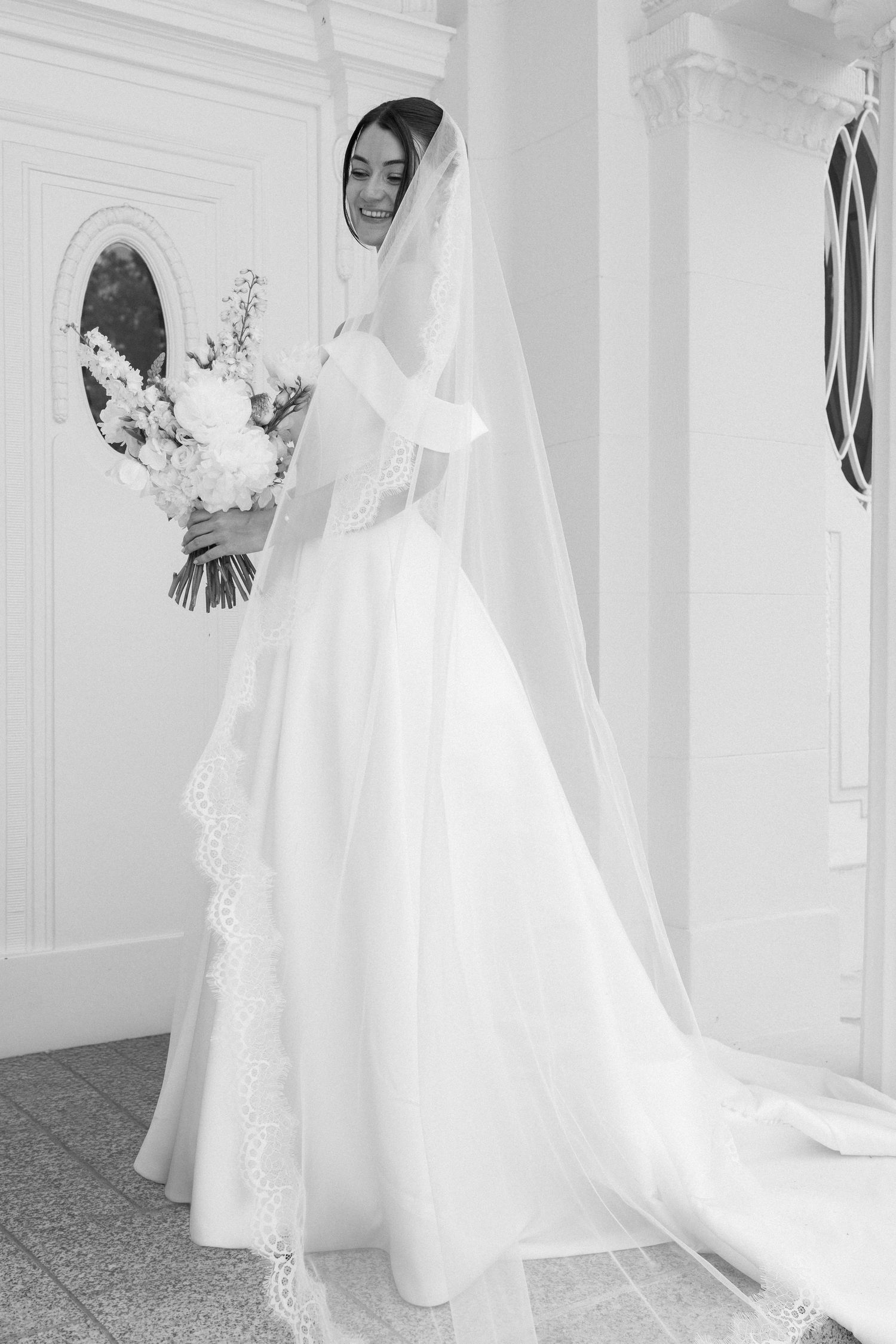 Elegant bride in white ball gown and lace veil holding floral bouquet, black and white wedding portrait.