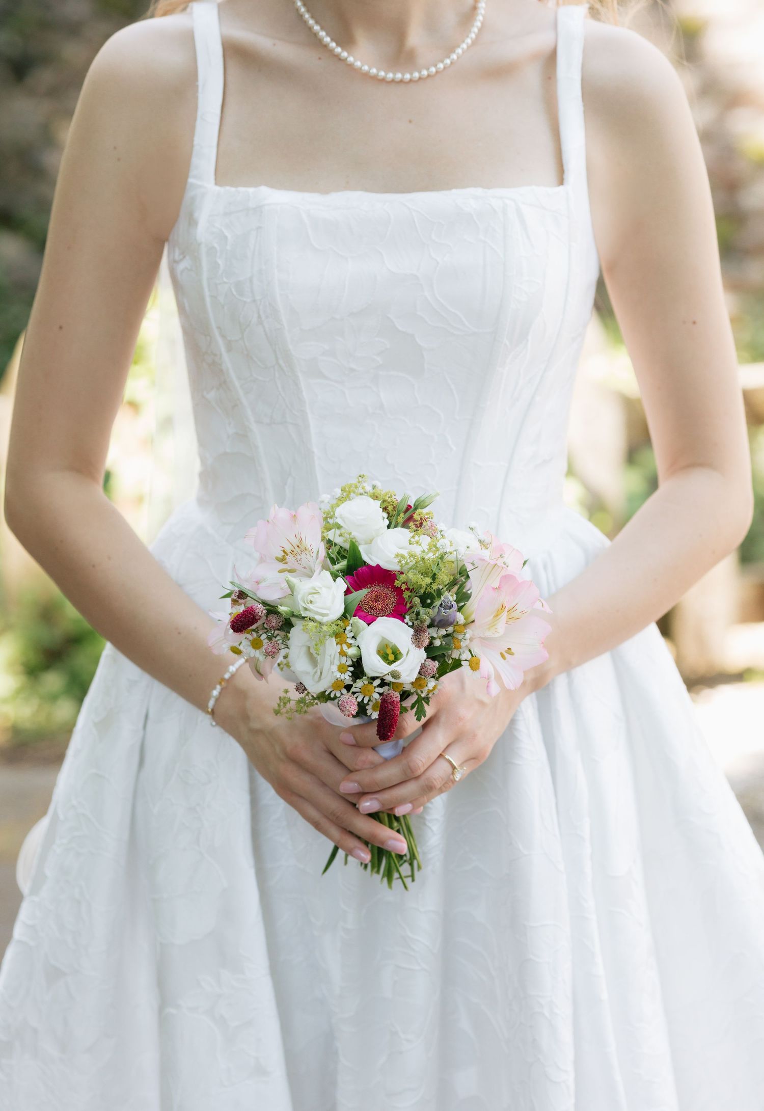 Bride in white square-neck wedding dress holding a small bouquet of pink, red, and white flowers with greenery.
