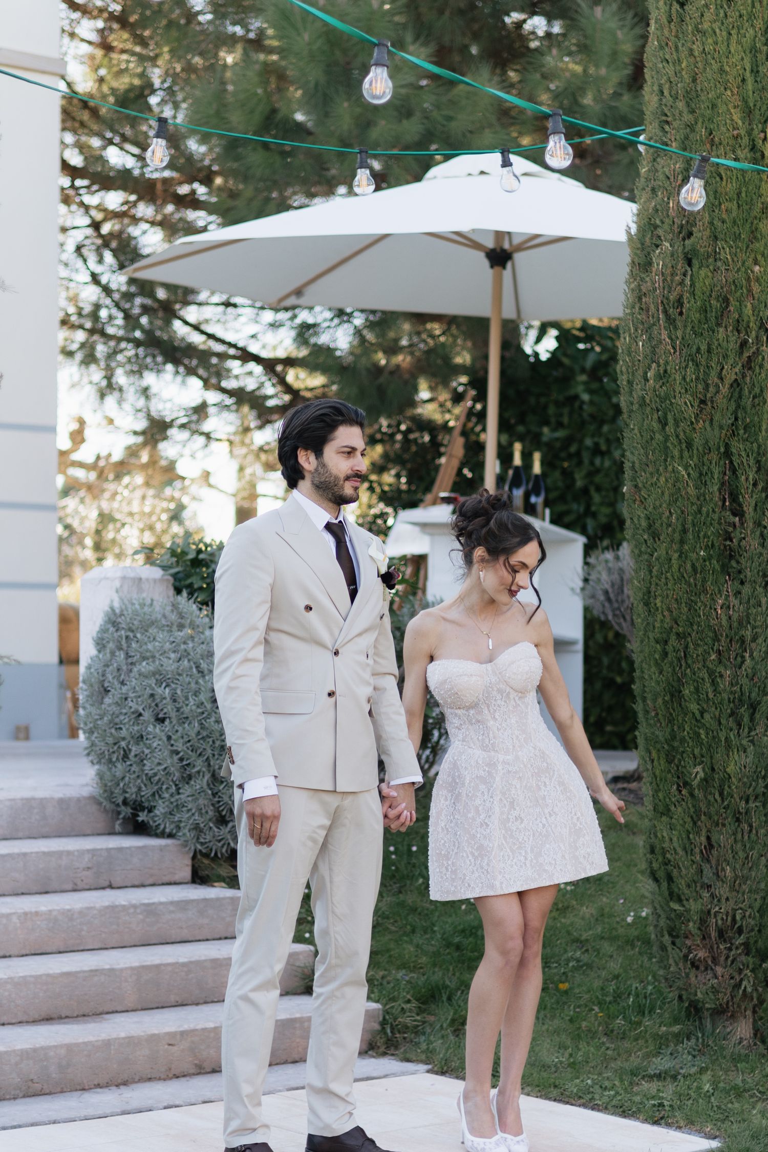 Elegant couple in wedding attire holding hands outdoors, groom in beige suit, bride in short lace dress, garden setting.