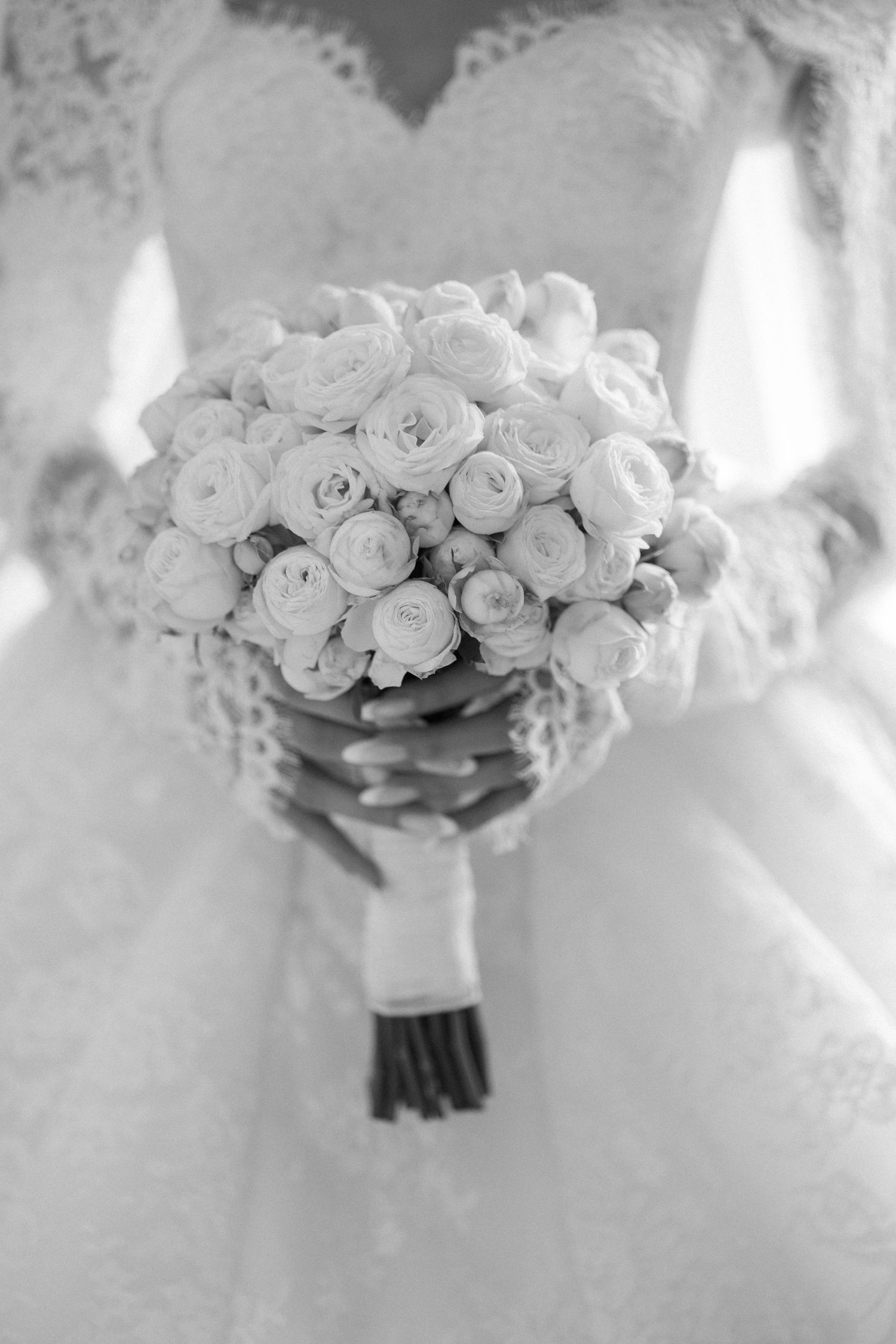 Bride in lace wedding dress holding a round bouquet of white roses in black and white photo.
