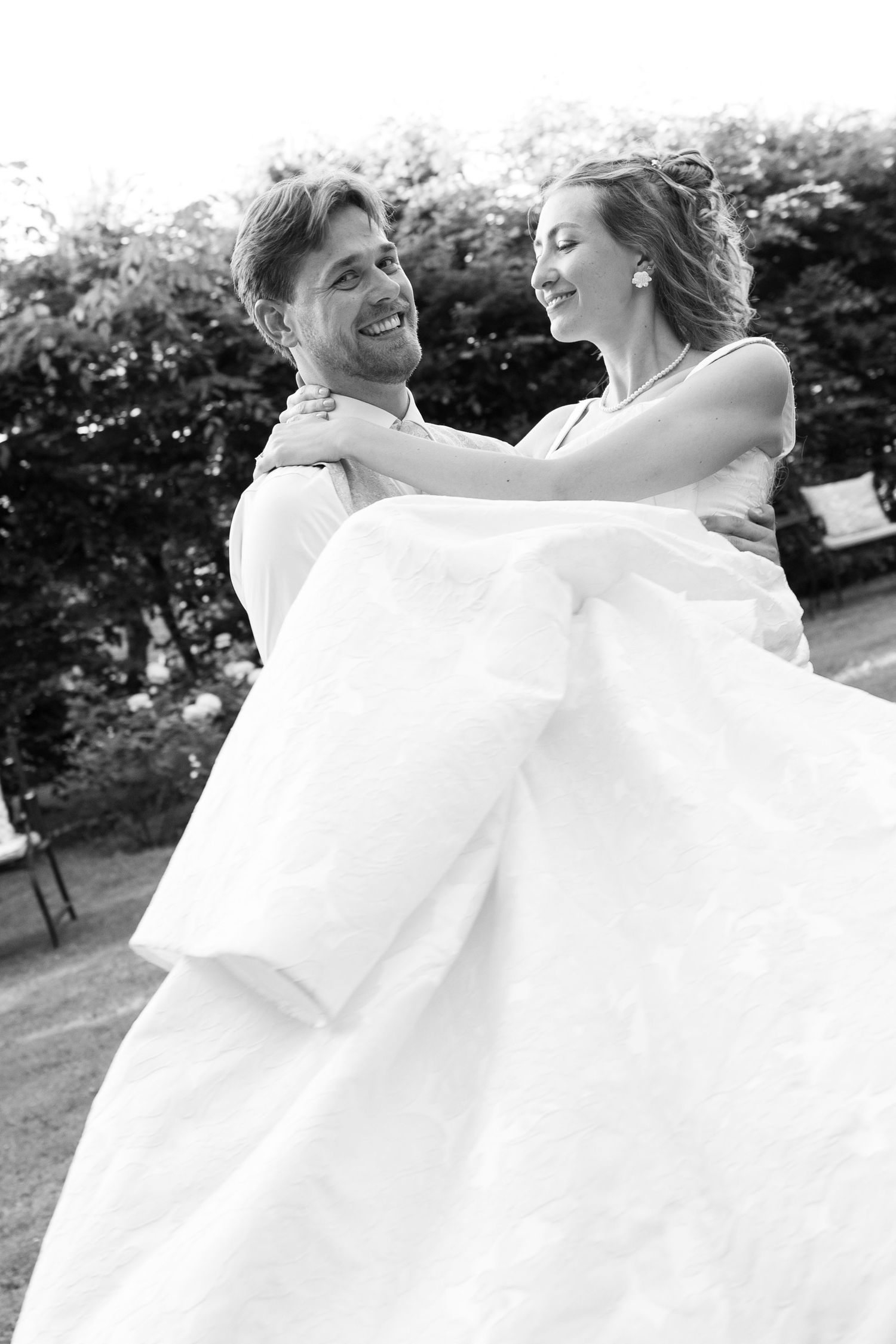 Groom carries bride in white wedding dress outdoors, both smiling joyfully, black and white photo.