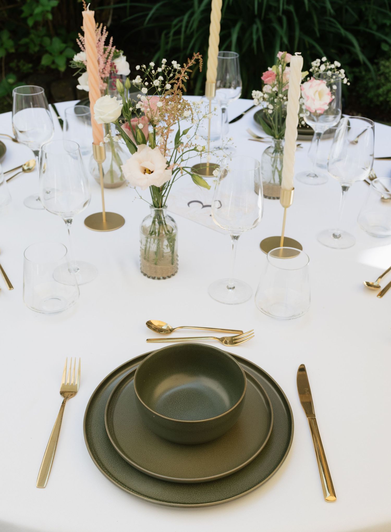 Elegant wedding table setting with olive green plates, gold cutlery, and floral centerpieces in glass vases on white linen.