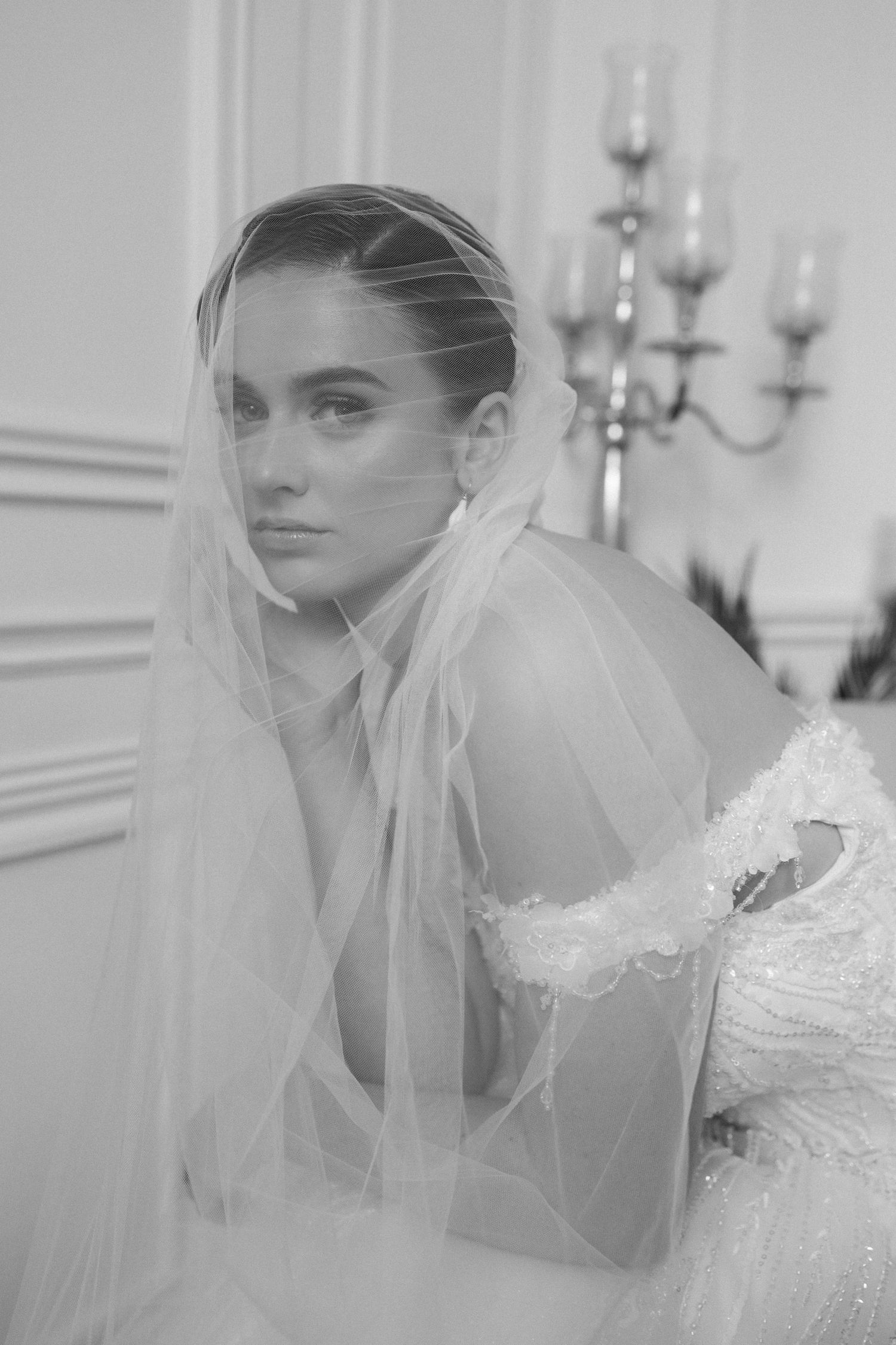 Black and white portrait of a bride wearing a veil and embellished gown, seated near a candelabra.