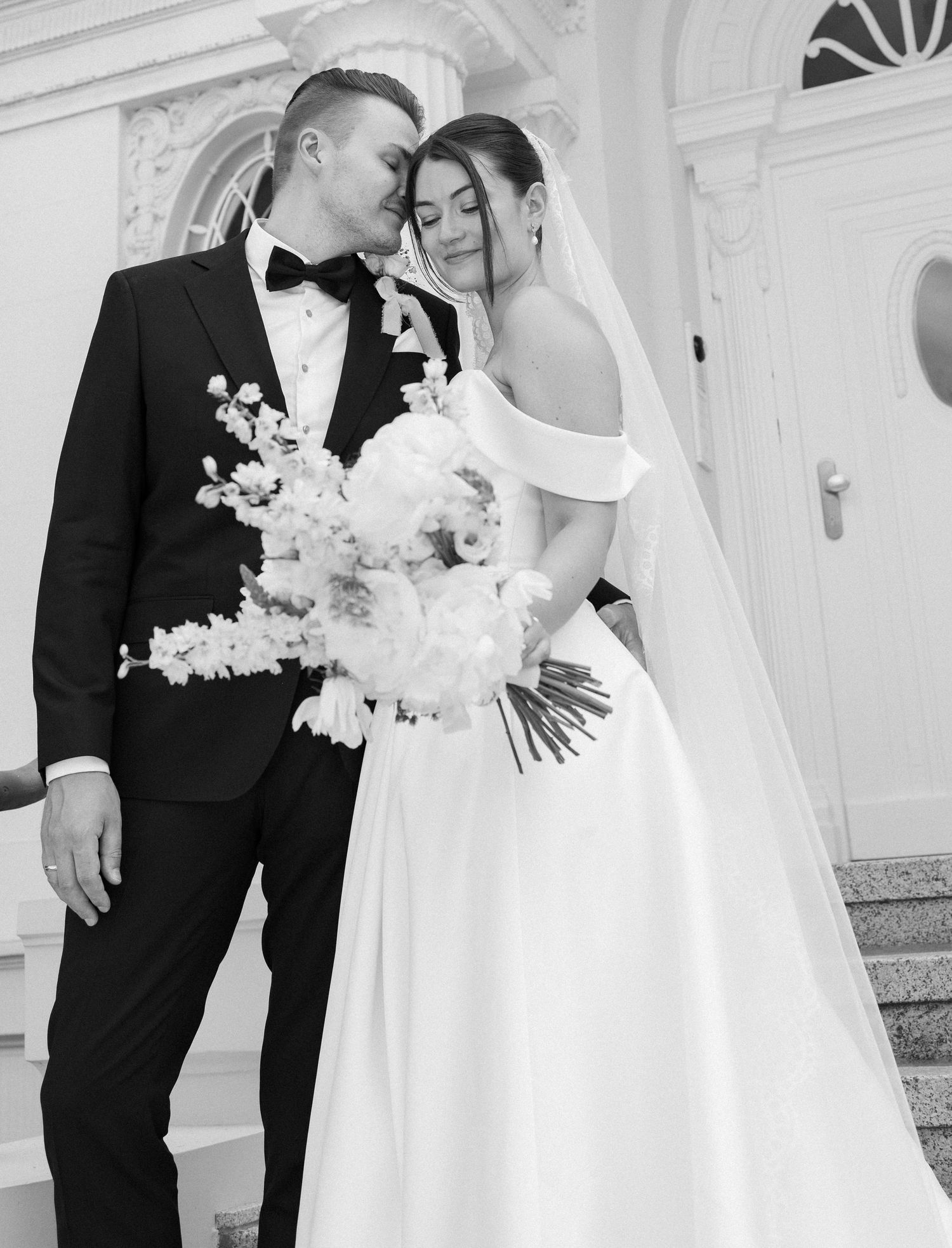 Black and white wedding photo of bride in off-shoulder gown holding bouquet, groom in tuxedo, sharing tender moment.