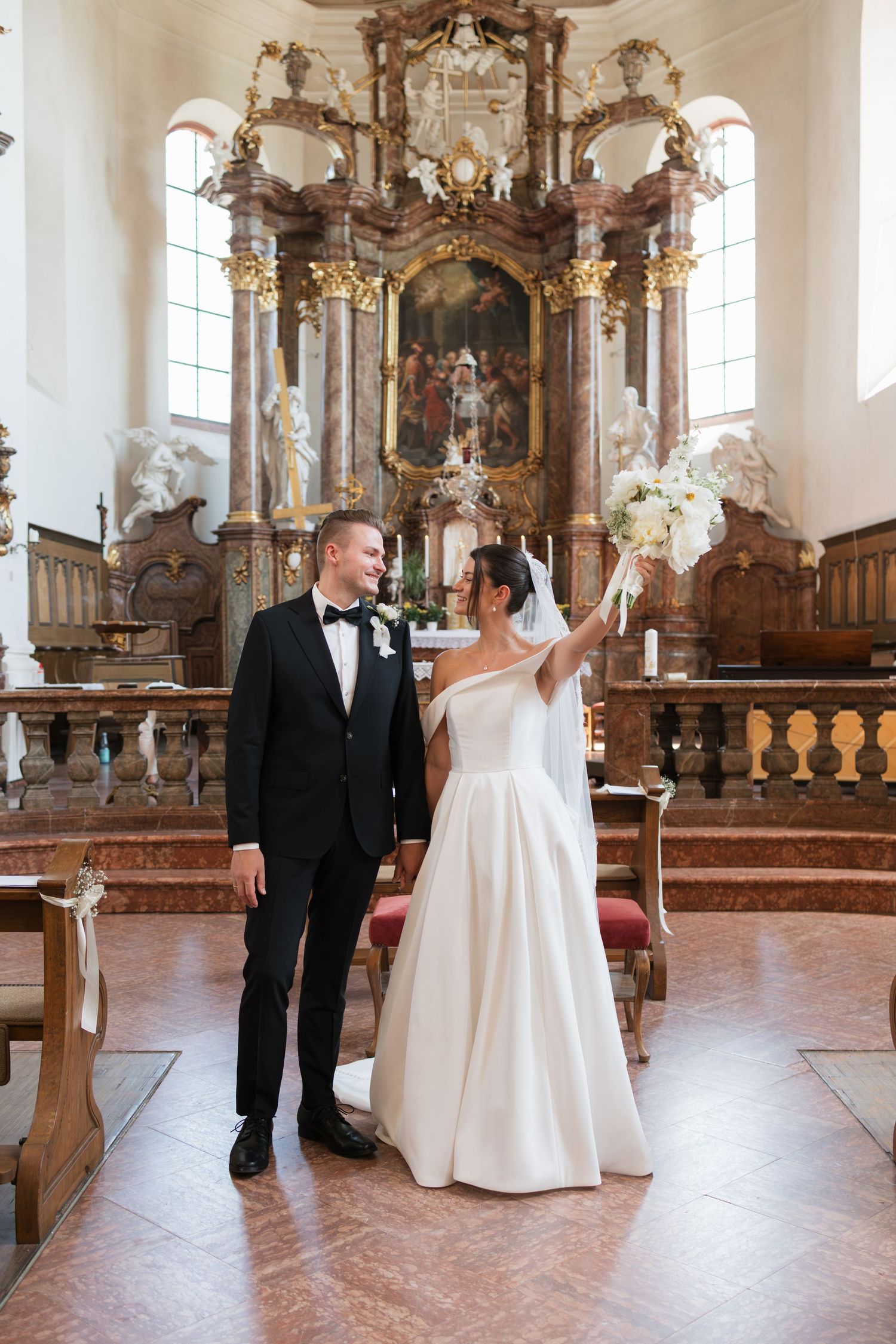 Bride and groom stand together in an ornate baroque church, bride raising white bouquet, groom in black tuxedo.