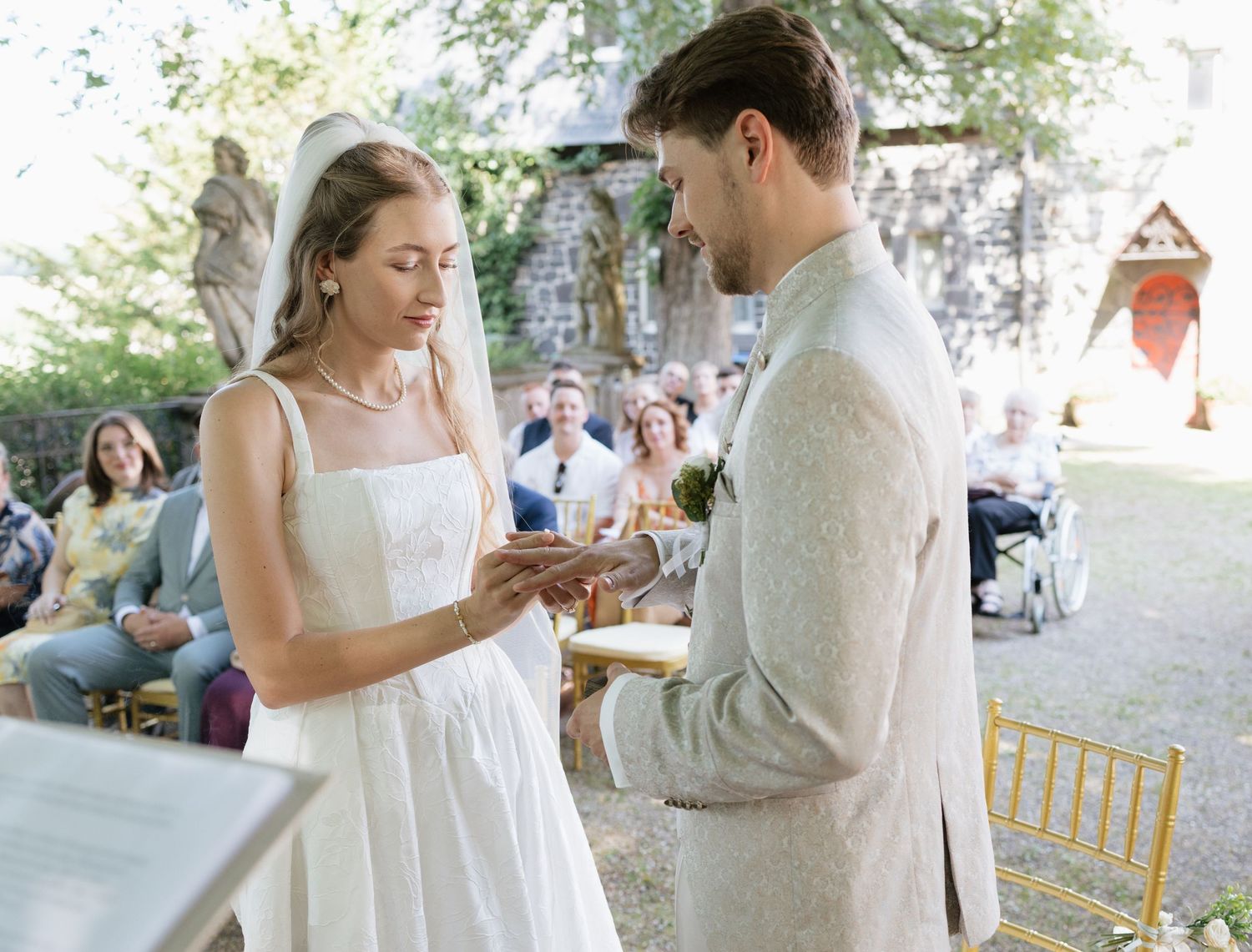 Couple exchanging rings at an outdoor wedding ceremony with guests seated in background.