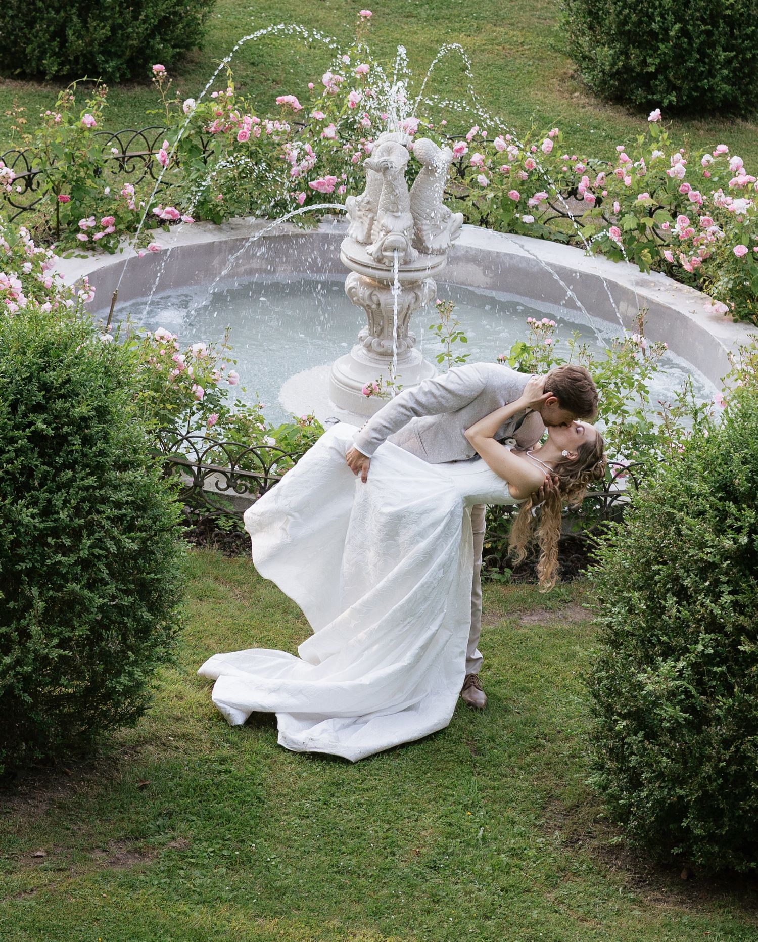 Bride and groom share a romantic dip kiss in front of a garden fountain surrounded by pink roses.