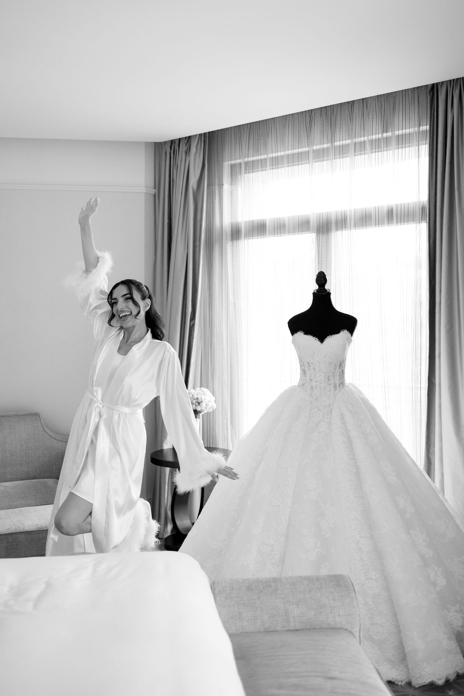 Black and white photo of a joyful bride in a robe celebrating next to her elegant ball gown wedding dress on a mannequin.