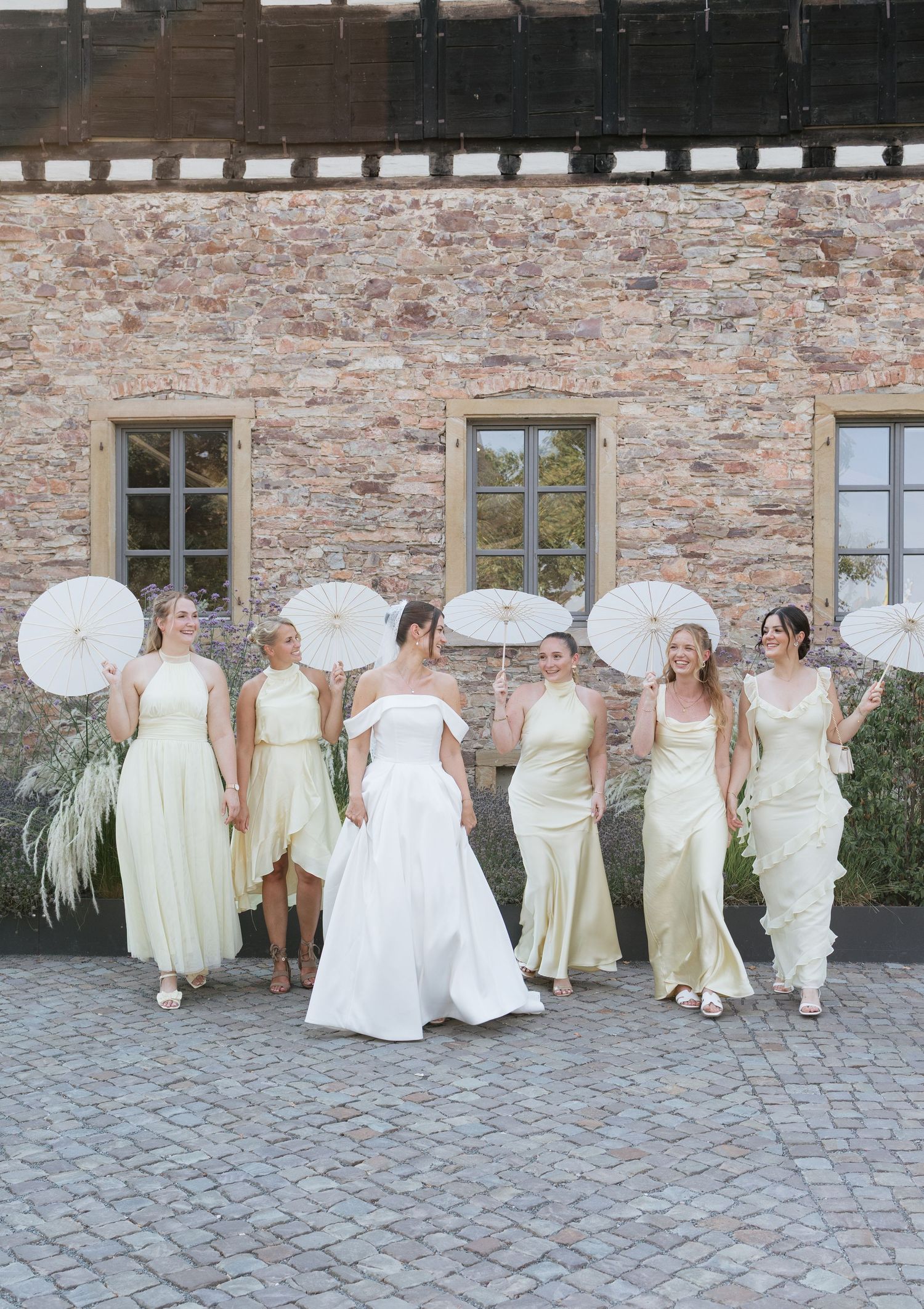 Bride in white gown poses with five bridesmaids in sage green dresses holding white parasols outside a stone building.