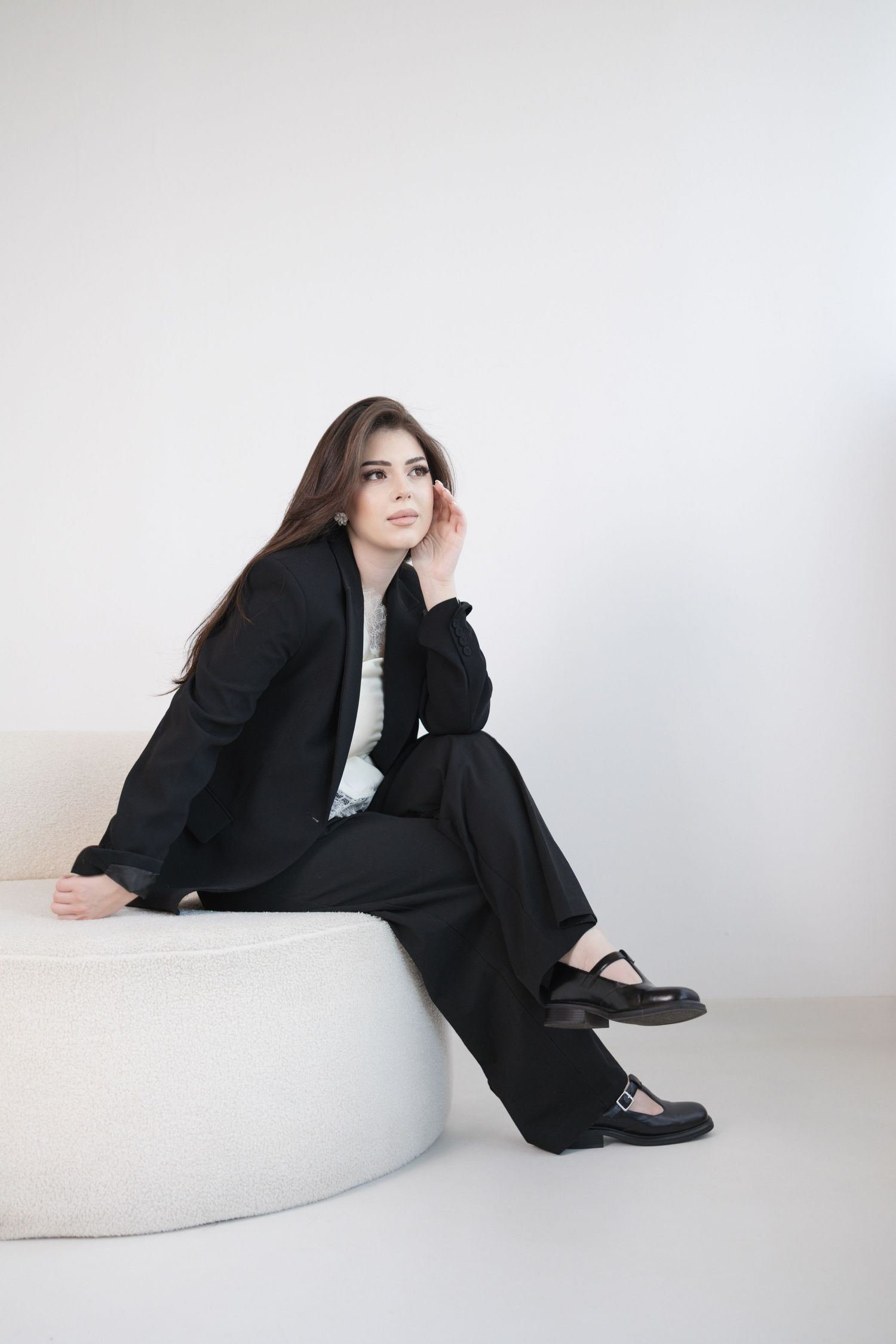 Young woman in black suit sitting on white round bench, posing thoughtfully in a minimalist studio setting.