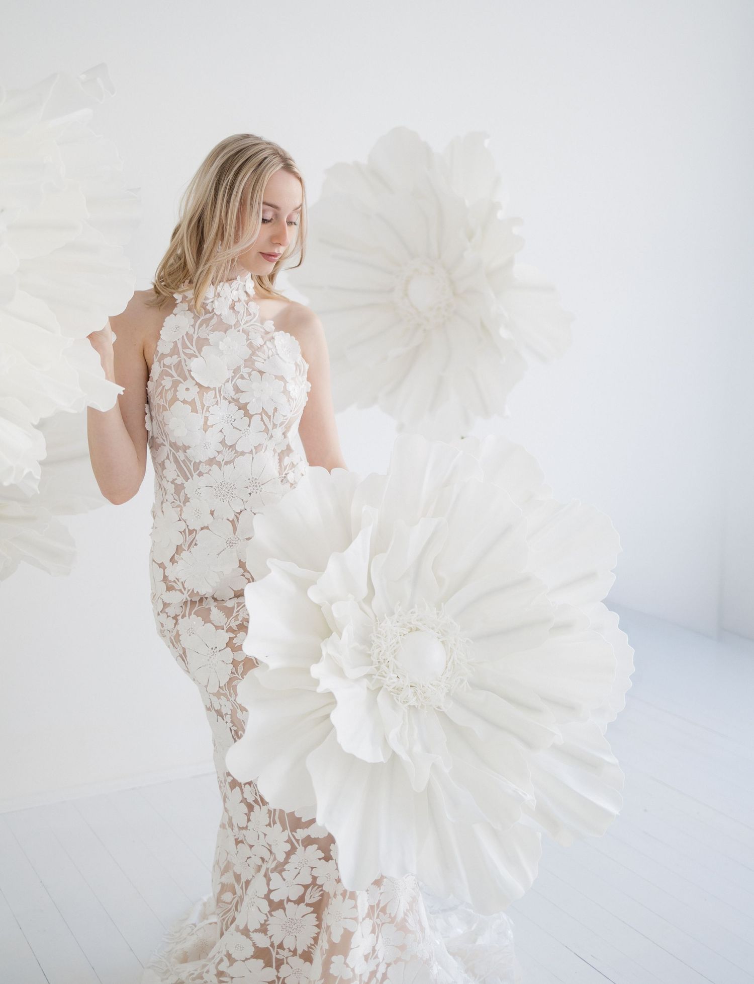 Bride in white lace halter gown poses among oversized white paper flowers in a bright studio setting.
