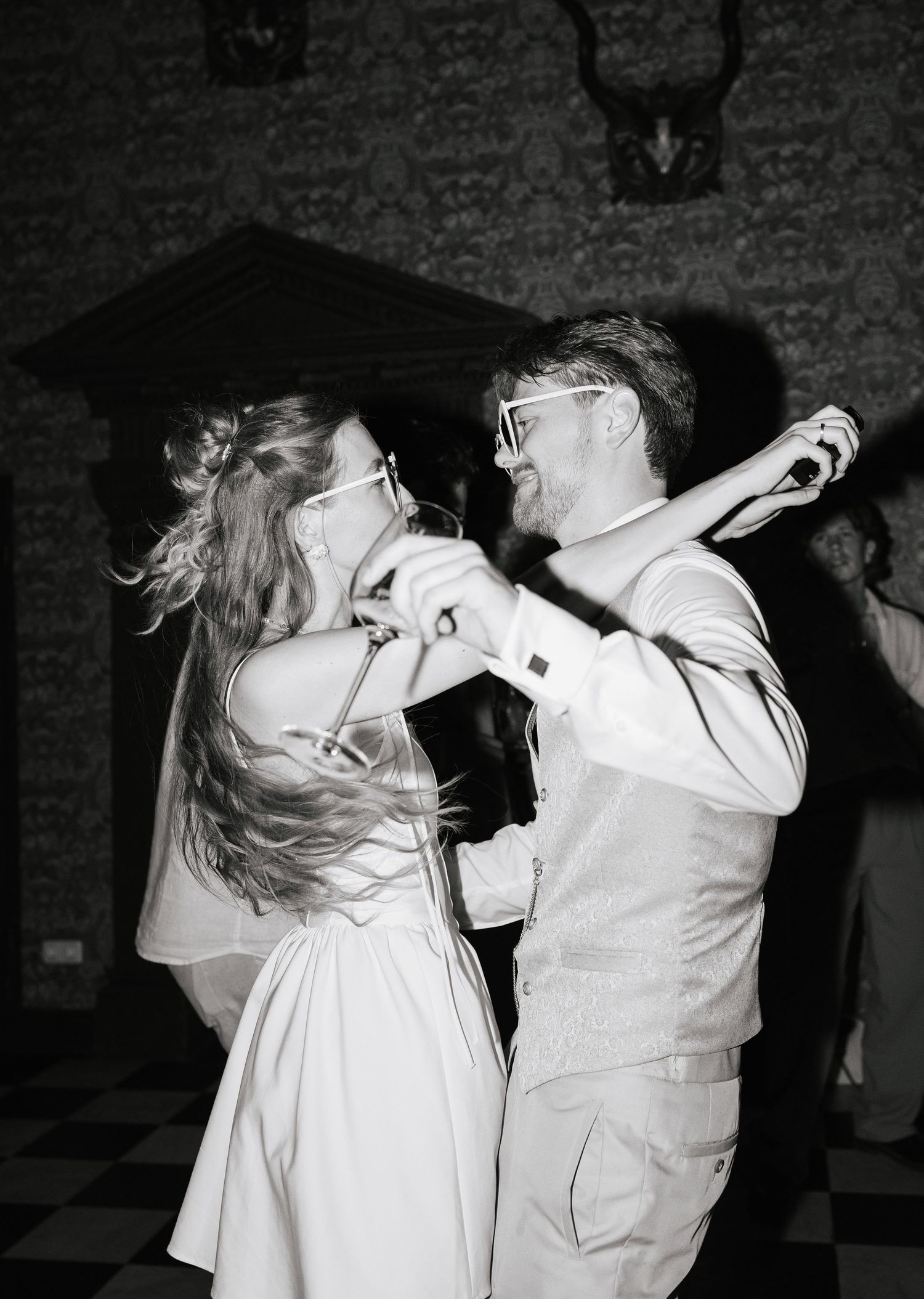 Bride and groom dancing joyfully at wedding reception in black and white photo, both wearing glasses.