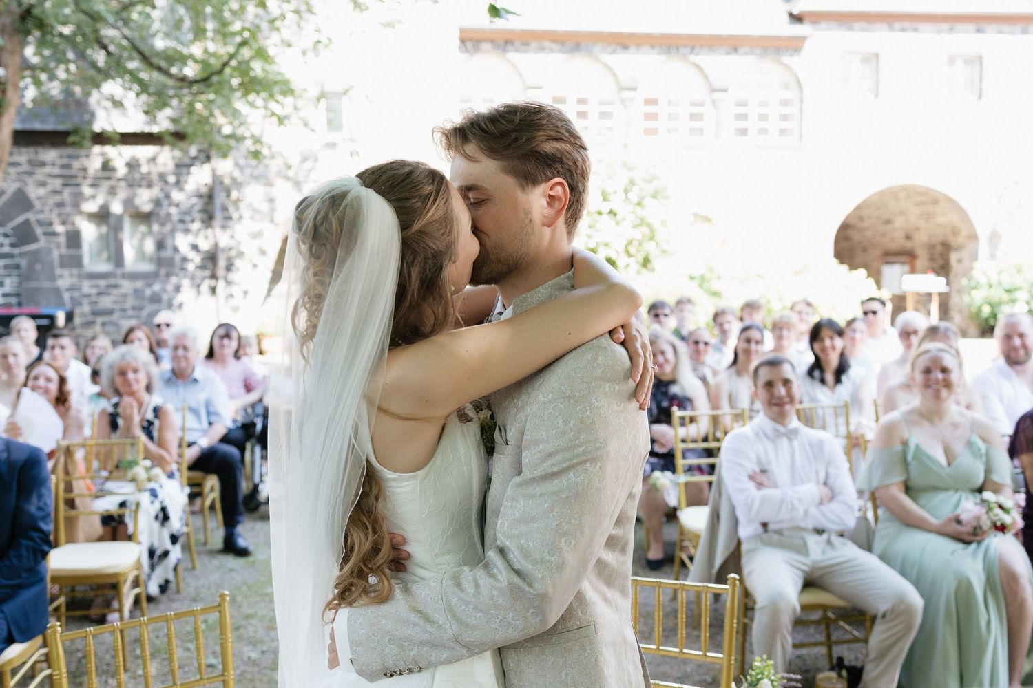Bride and groom share first kiss at outdoor wedding ceremony with guests seated in gold chairs.