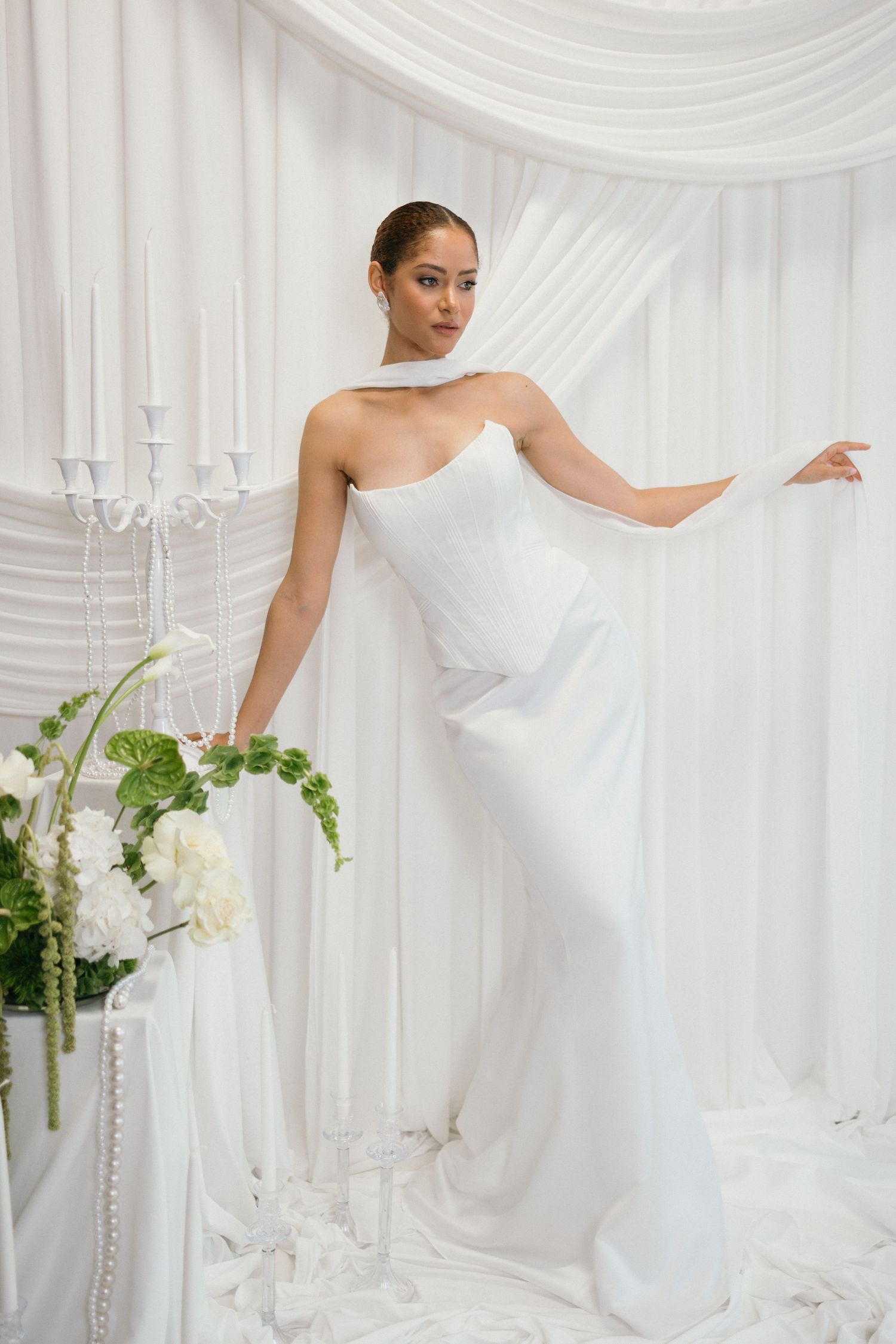 Elegant bride in a sleek white one-shoulder gown poses gracefully among white floral arrangements and draped fabric backdrop.