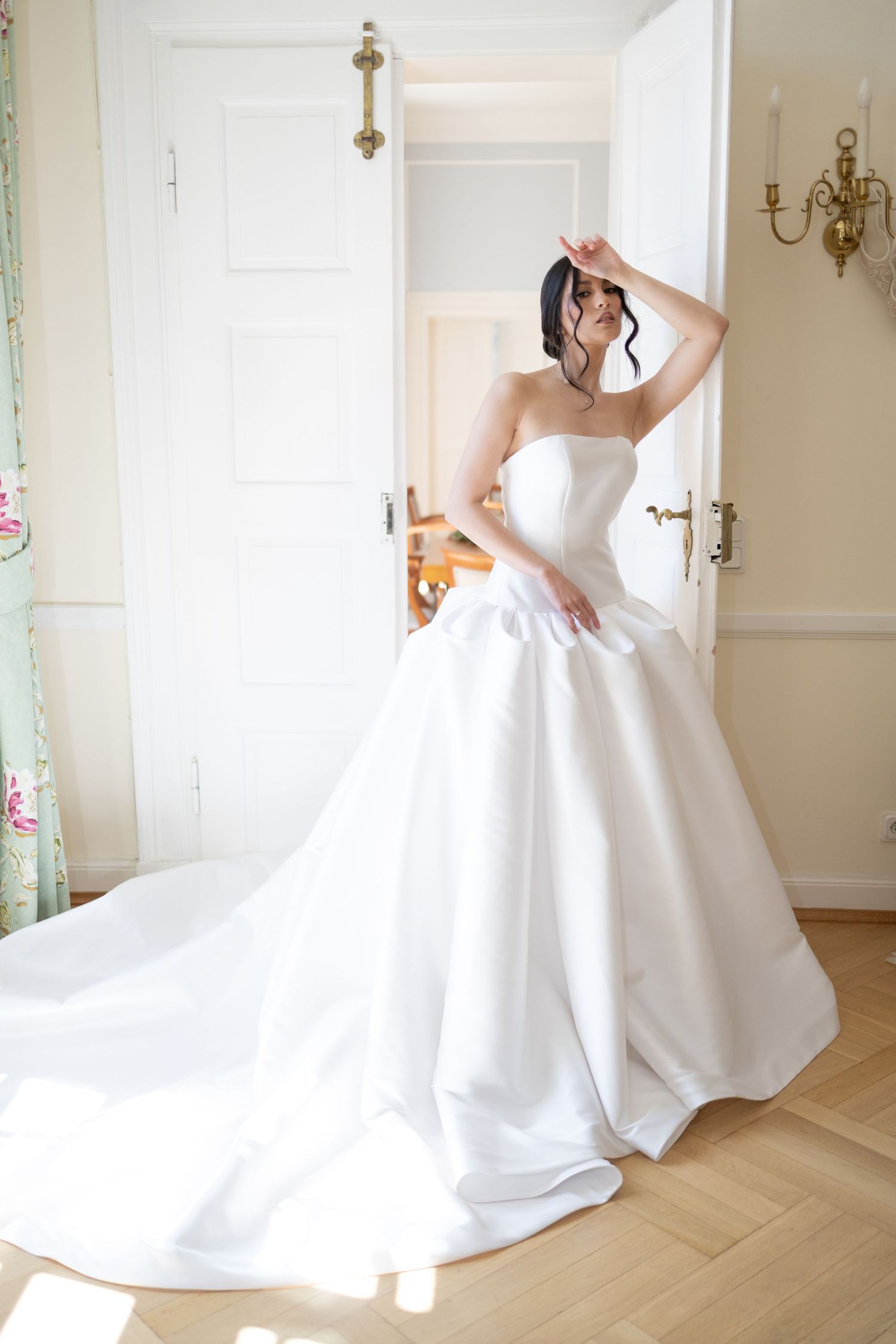 Bride in a strapless white ball gown with a long train stands by an open door in a sunlit elegant room.