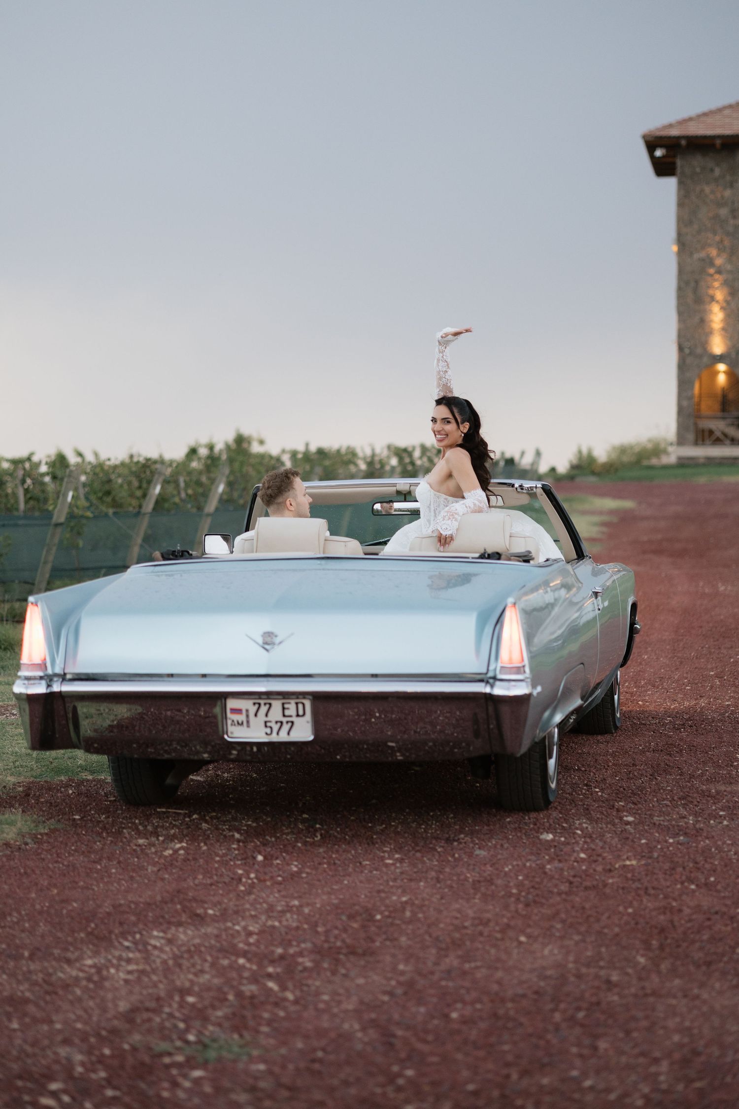 Bride in white dress waves from a classic light blue convertible on a rustic dirt road at dusk.