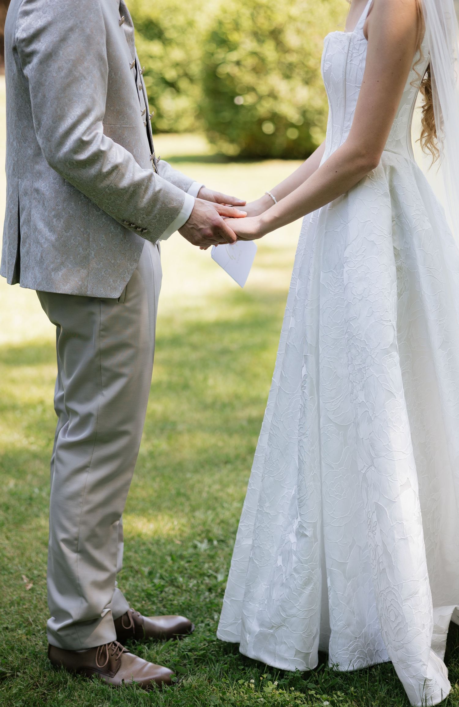 Bride and groom holding hands outdoors during wedding ceremony, groom in gray suit, bride in white lace gown.