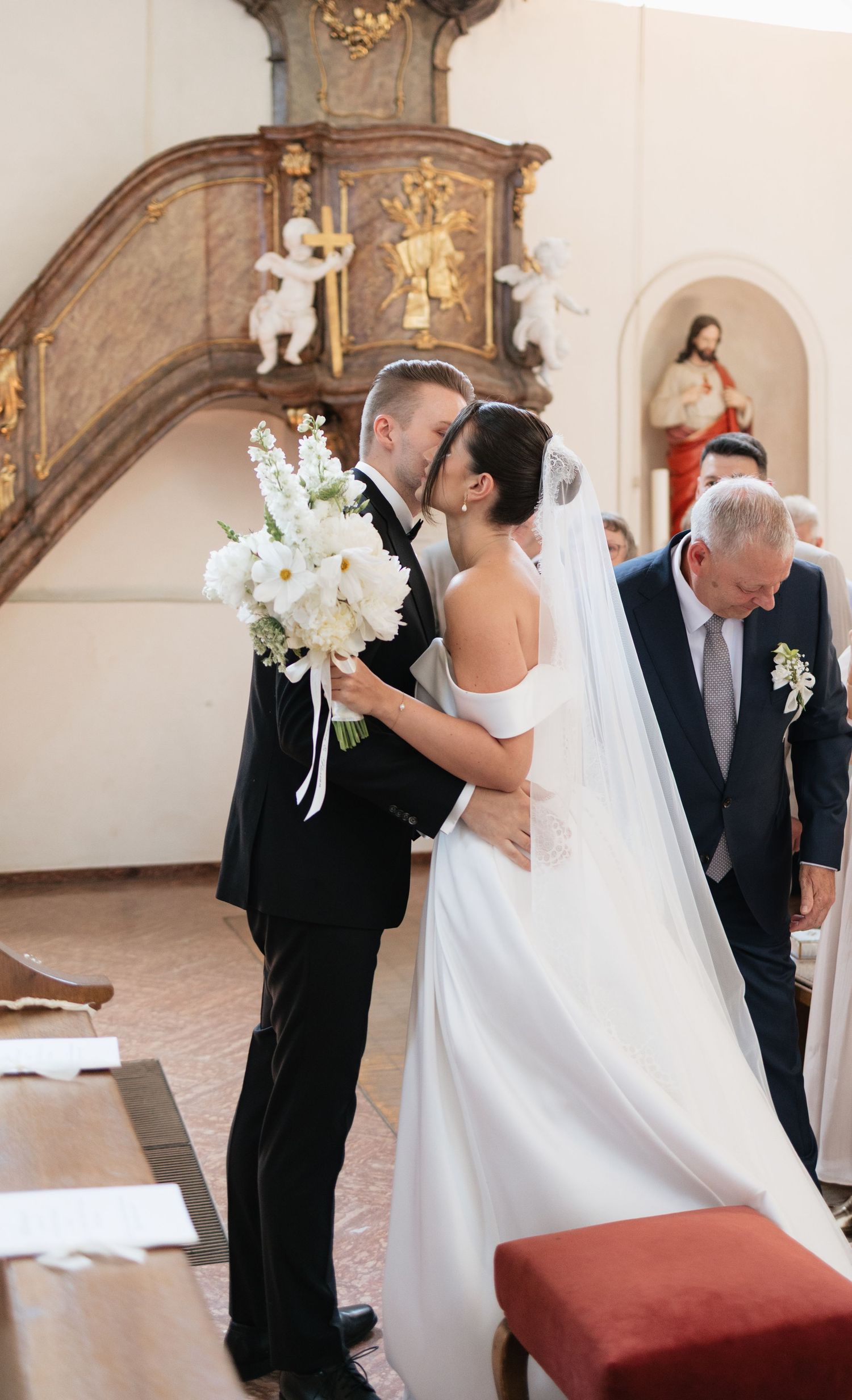 Bride and groom share a kiss at church altar, bride holding white floral bouquet in elegant off-shoulder gown.