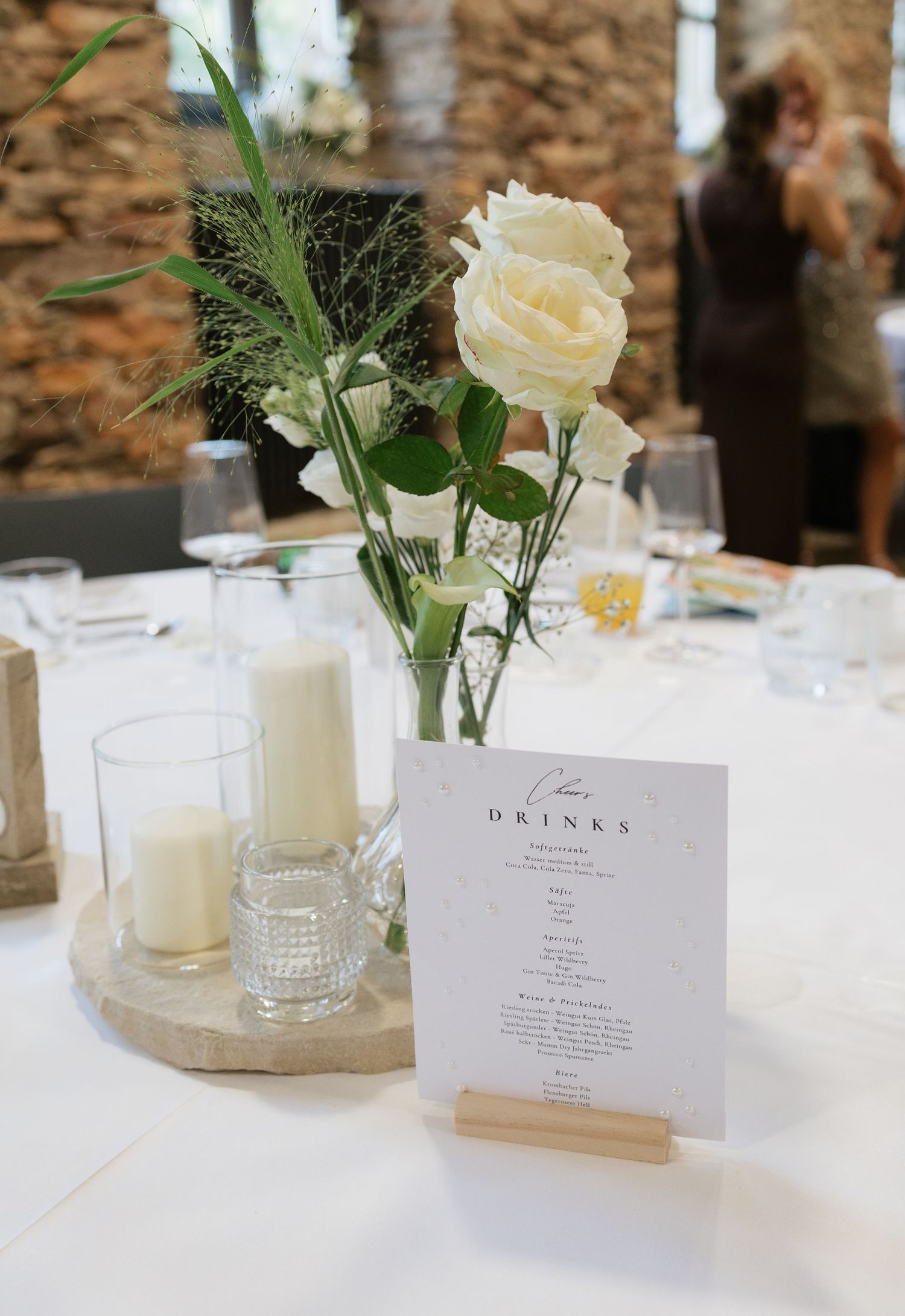 Elegant wedding table centerpiece with white roses, candle, and drinks menu card on a wooden slice.