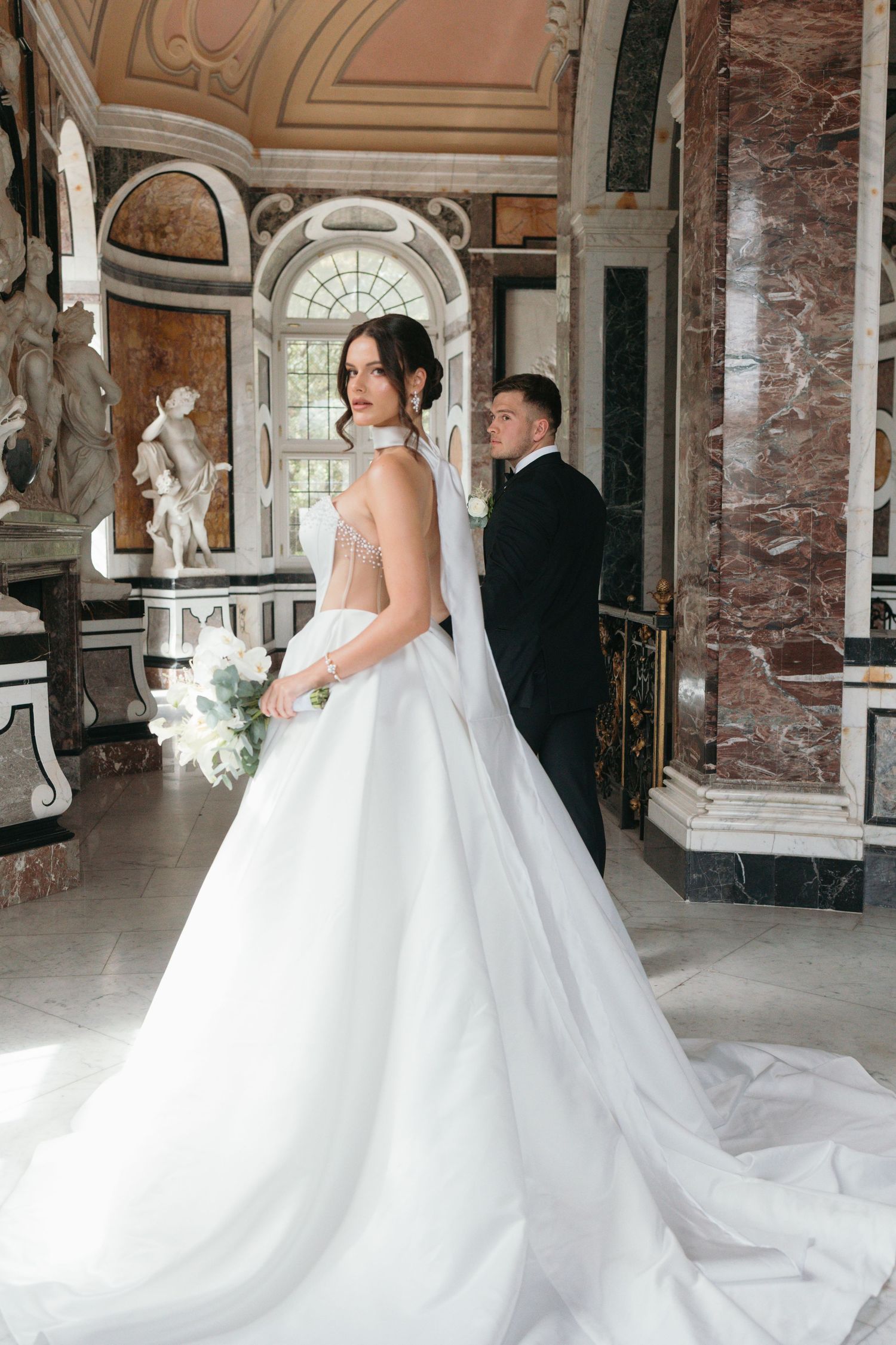 Elegant bride in a flowing white gown holds a bouquet beside a tuxedo-clad groom in a grand marble hall.