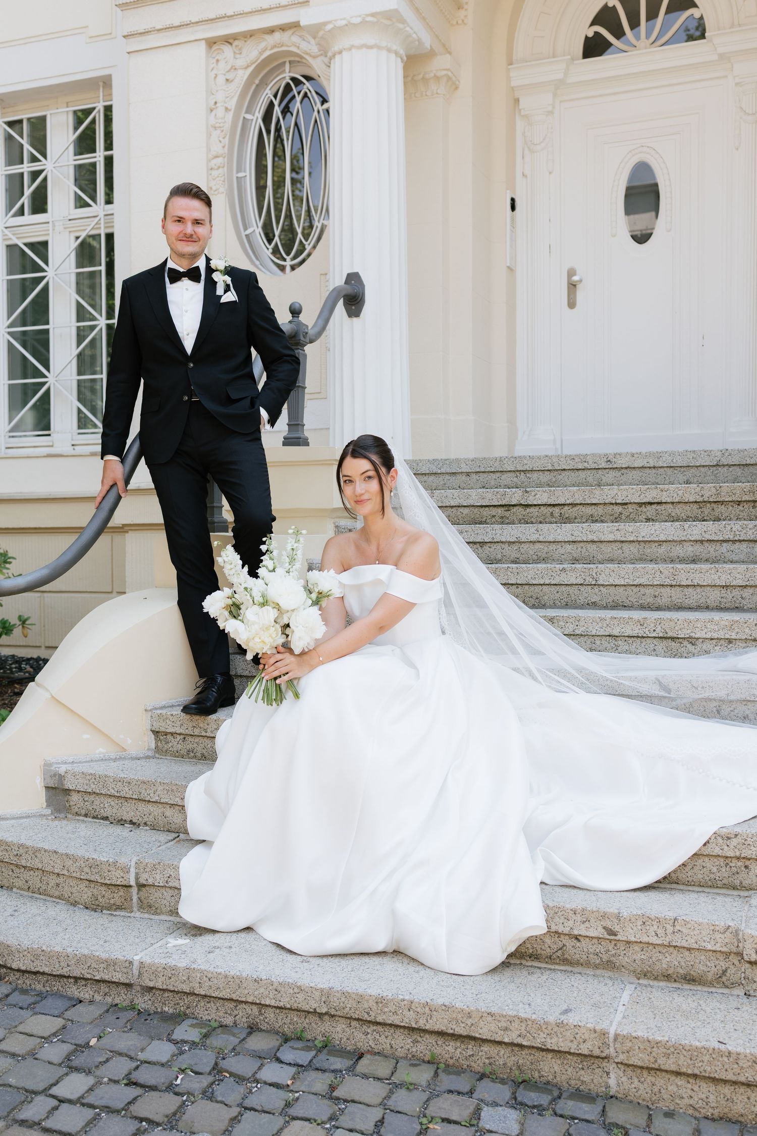 Bride in white off-shoulder gown sits on steps holding bouquet while groom in black tuxedo stands behind her.