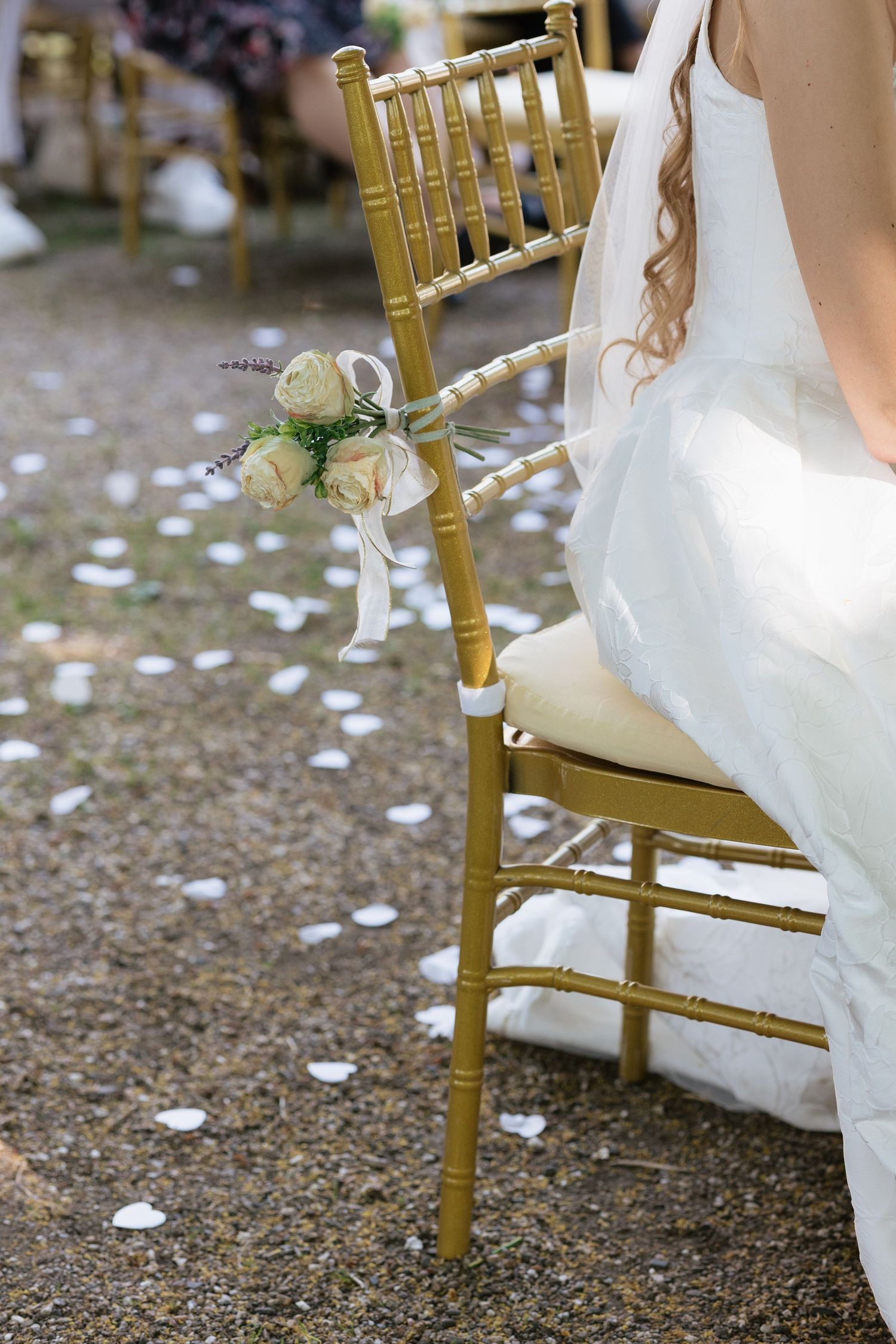 Gold Chiavari chair with white rose bouquet at outdoor wedding ceremony, white petals scattered on ground.