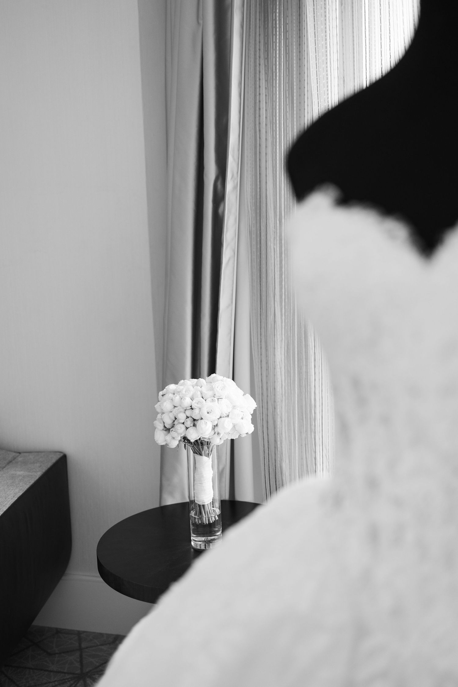Black and white photo of a white floral bouquet in a glass vase on a table beside sheer curtains and a wedding dress.
