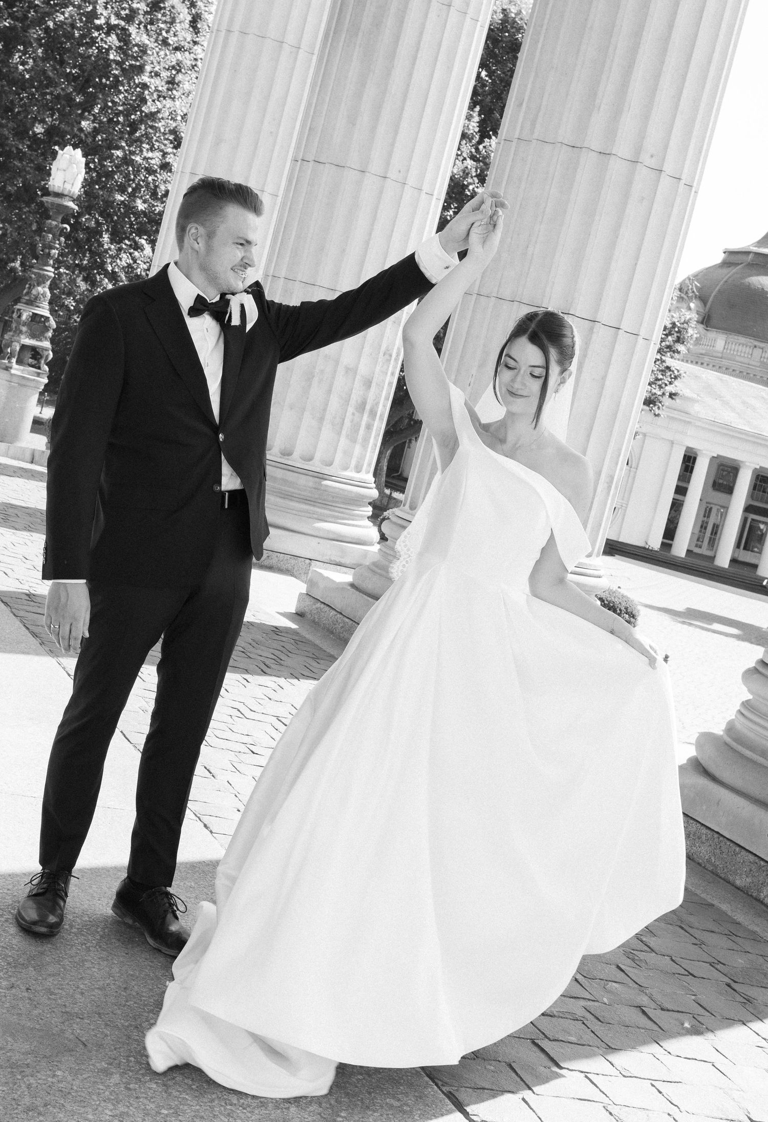 Black and white photo of a groom spinning his bride in a white gown near elegant columns.