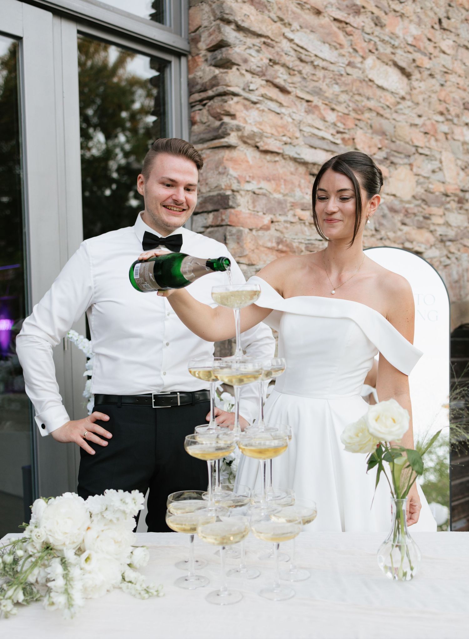 Bride and groom pouring champagne tower at wedding reception outdoors near stone wall.