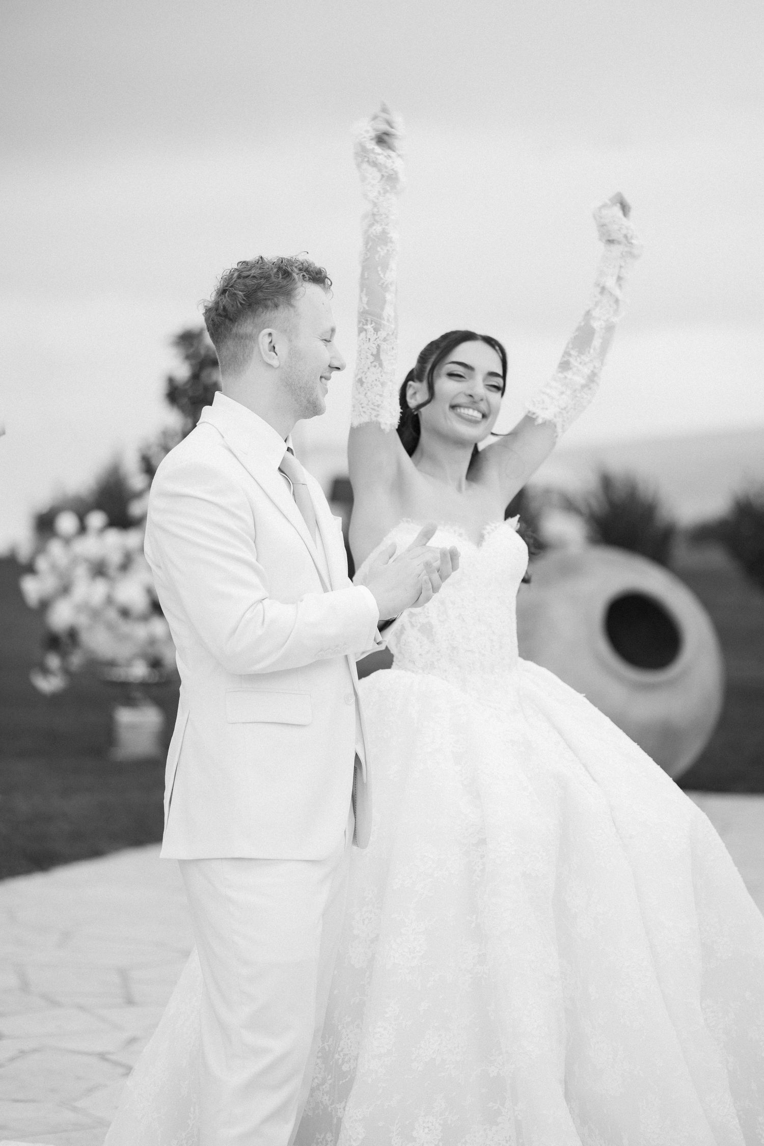 Black and white photo of a joyful bride in a ball gown raising her arms with a groom in a white suit outdoors.
