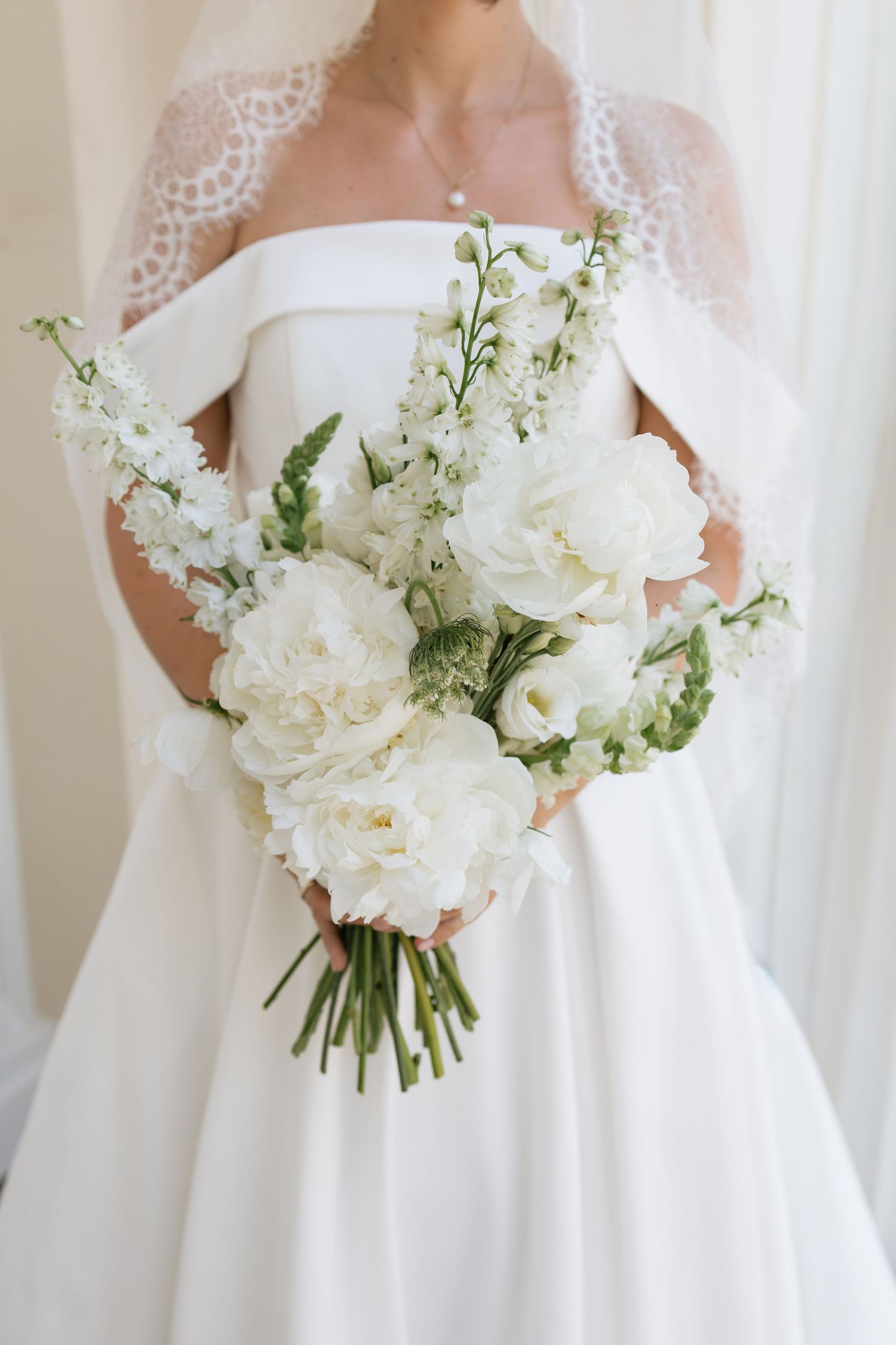 Bride in white off-shoulder gown holding elegant bouquet of white peonies and delphinium with lace veil.