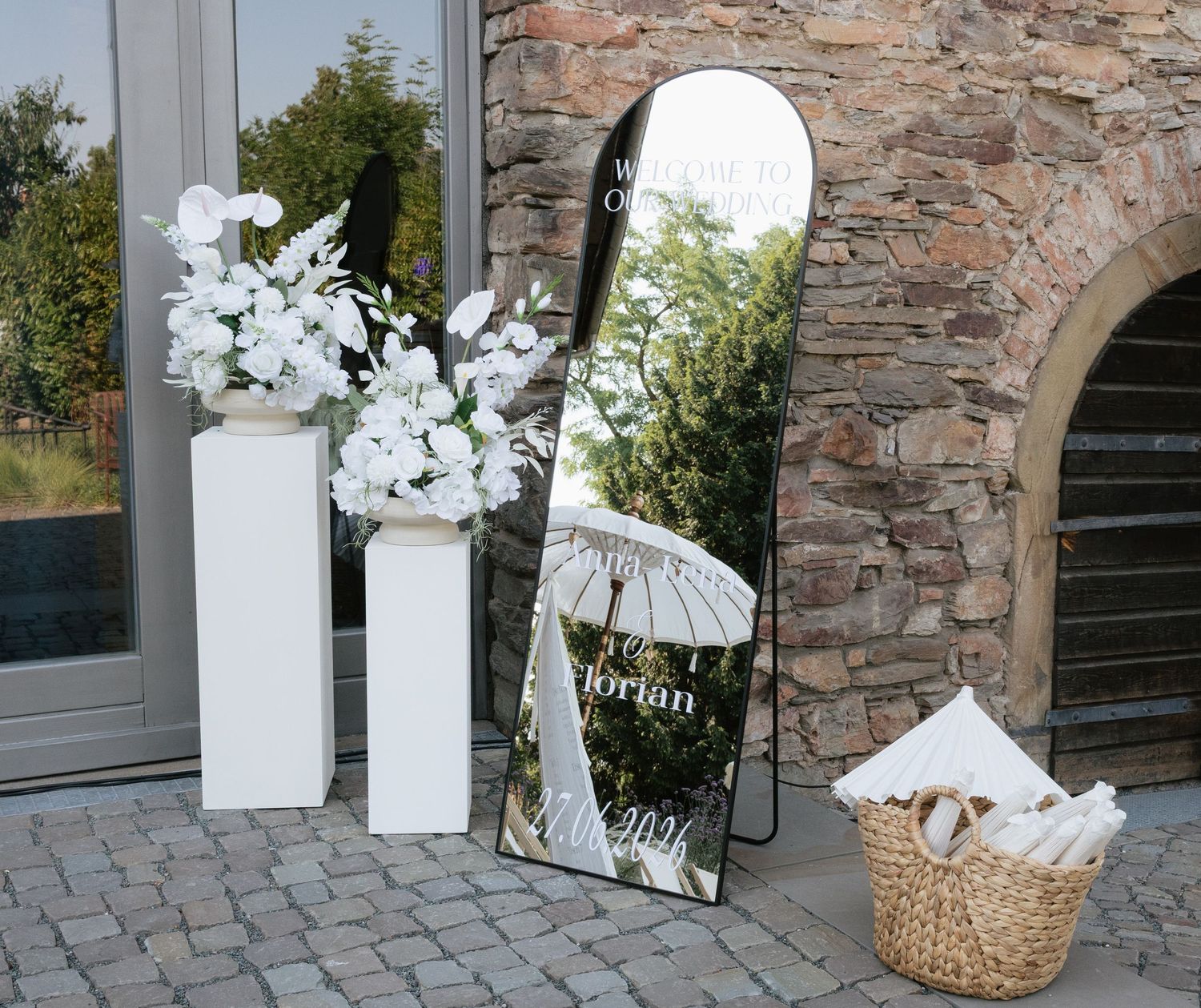 Elegant wedding mirror sign with white floral arrangements on pedestals and wicker basket outside stone venue.