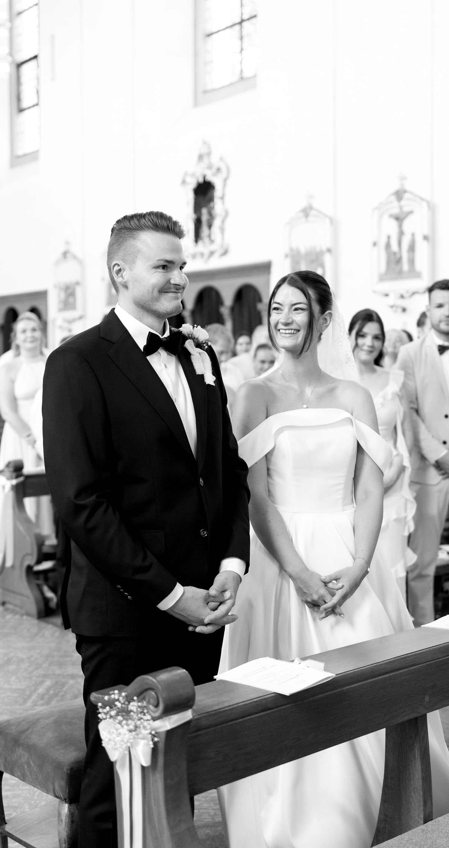 Black and white photo of smiling bride and groom holding hands at church altar during wedding ceremony.