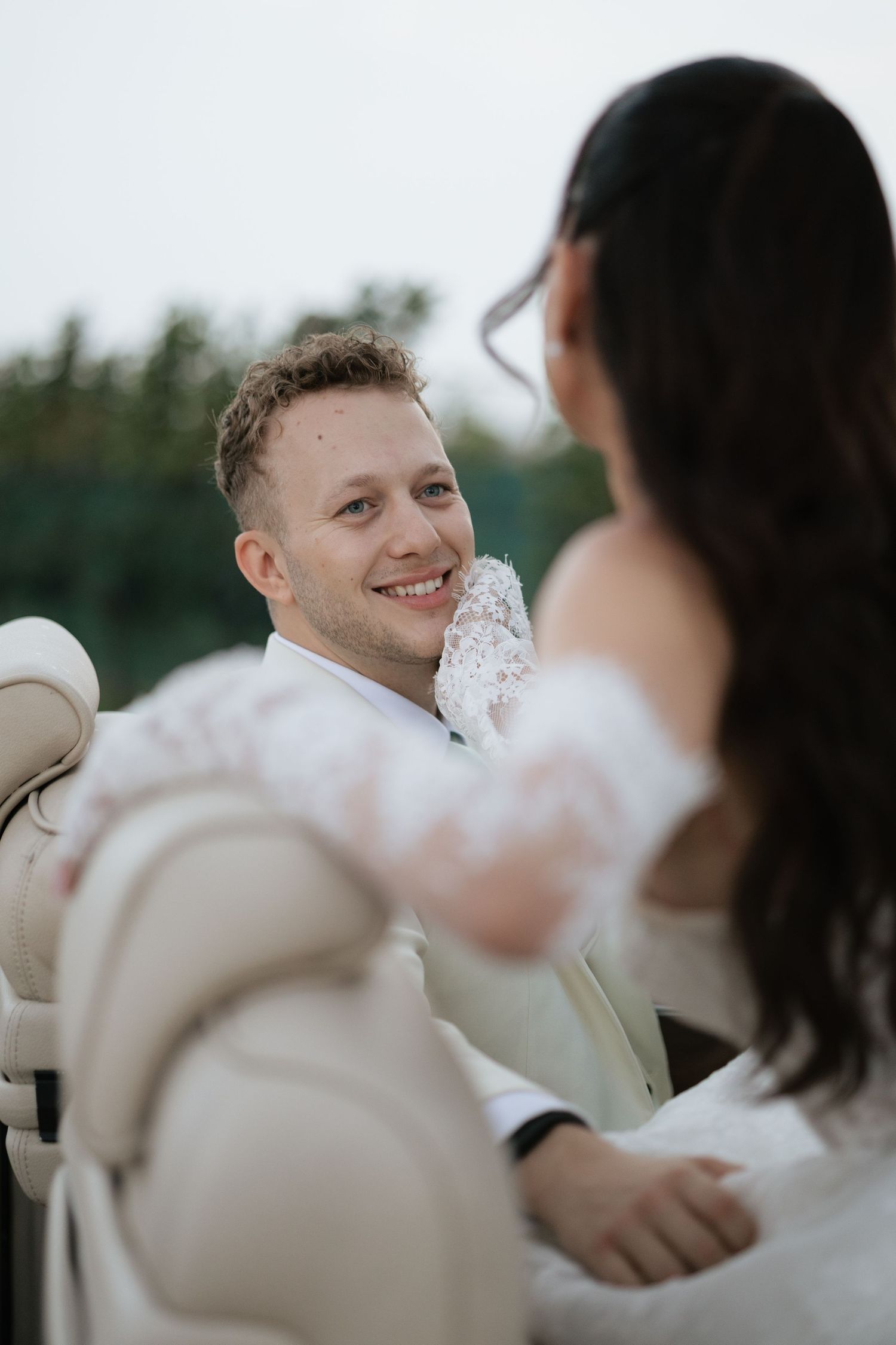 Smiling groom in white suit looks lovingly at bride in lace gloves during outdoor wedding ceremony.