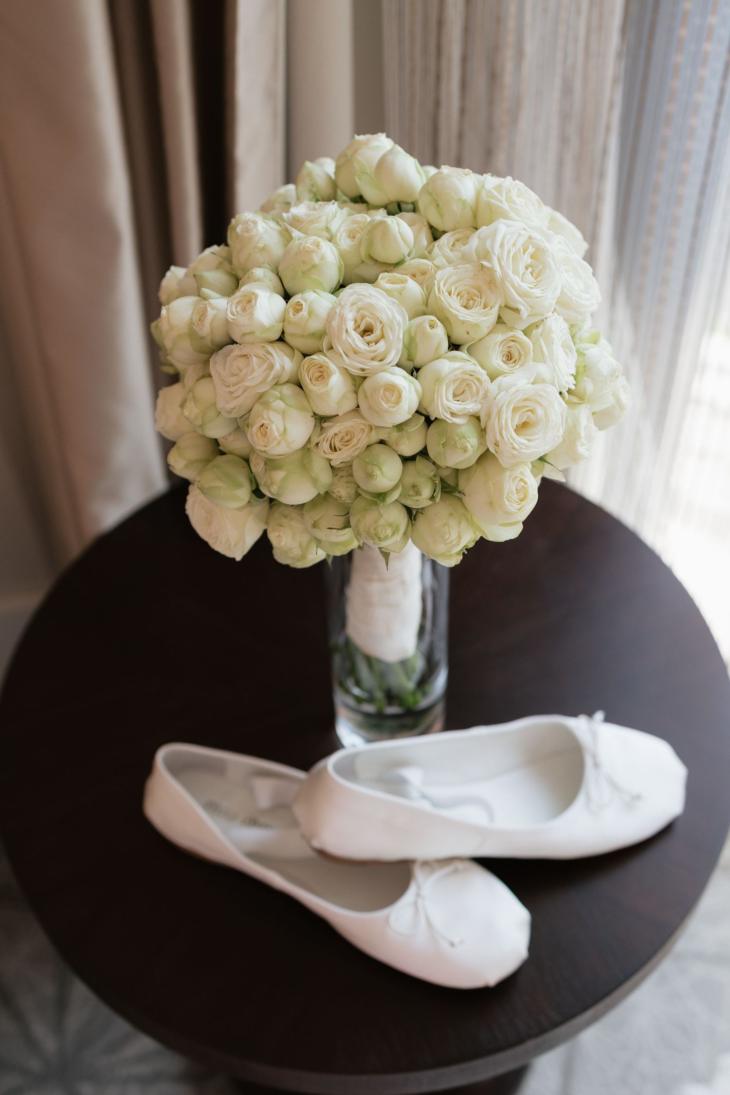 White bridal ballet flats with bow detail beside a lush white rose bouquet in a glass vase on a dark round table.