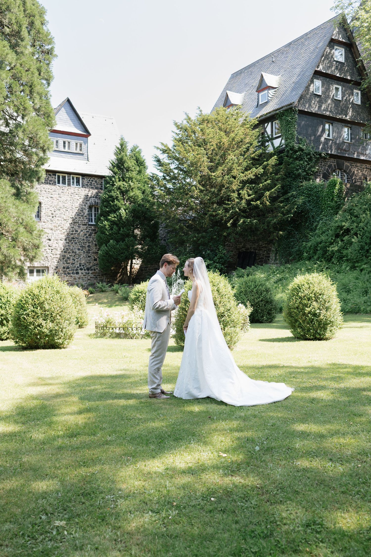 Bride and groom stand together on lush green lawn before a historic stone and timber manor house on their wedding day.