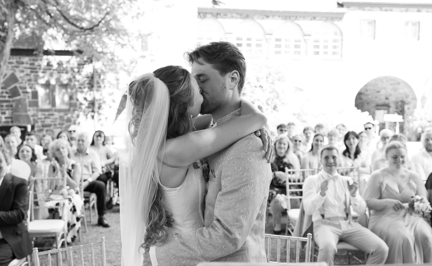 Bride and groom share first kiss at outdoor wedding ceremony with seated guests in black and white photo.