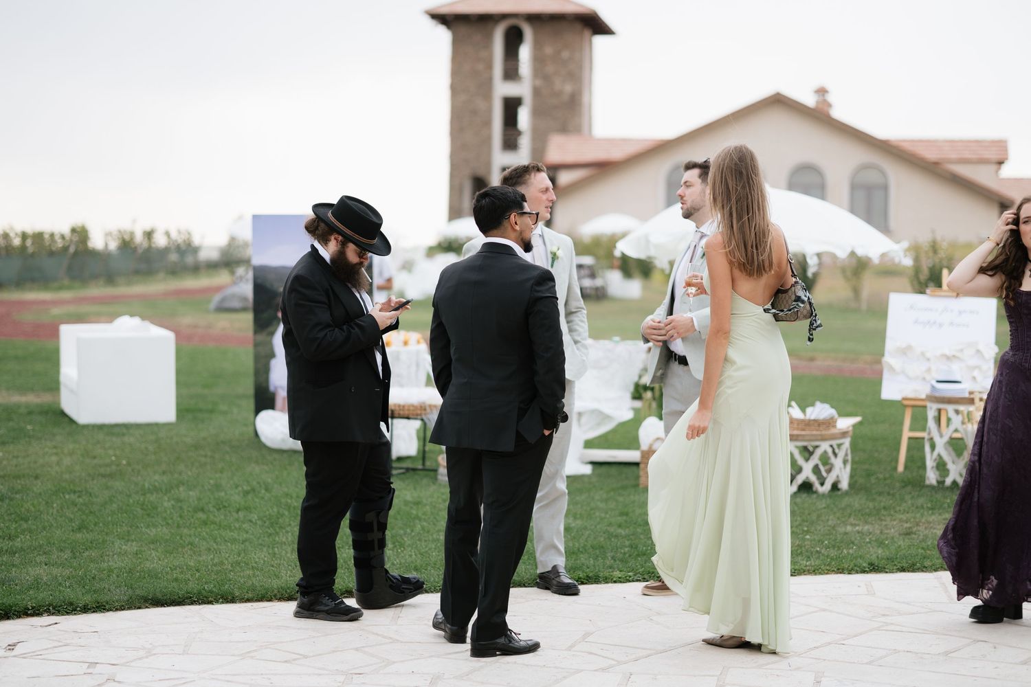 Guests in formal attire mingle outdoors at an elegant wedding reception on a green lawn near a stone building.