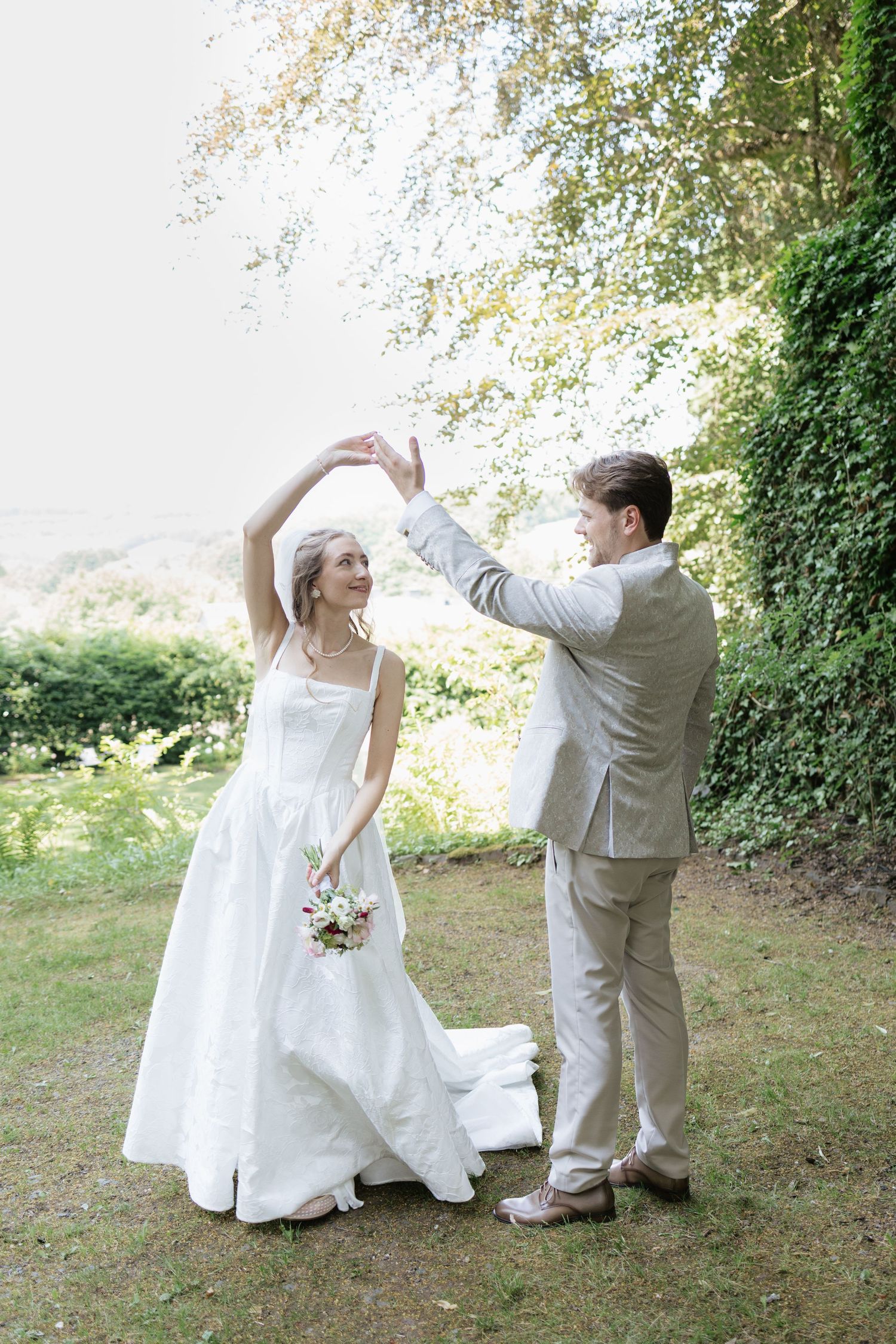 Bride and groom dancing outdoors in lush garden, bride in white gown holding bouquet, groom in beige suit.