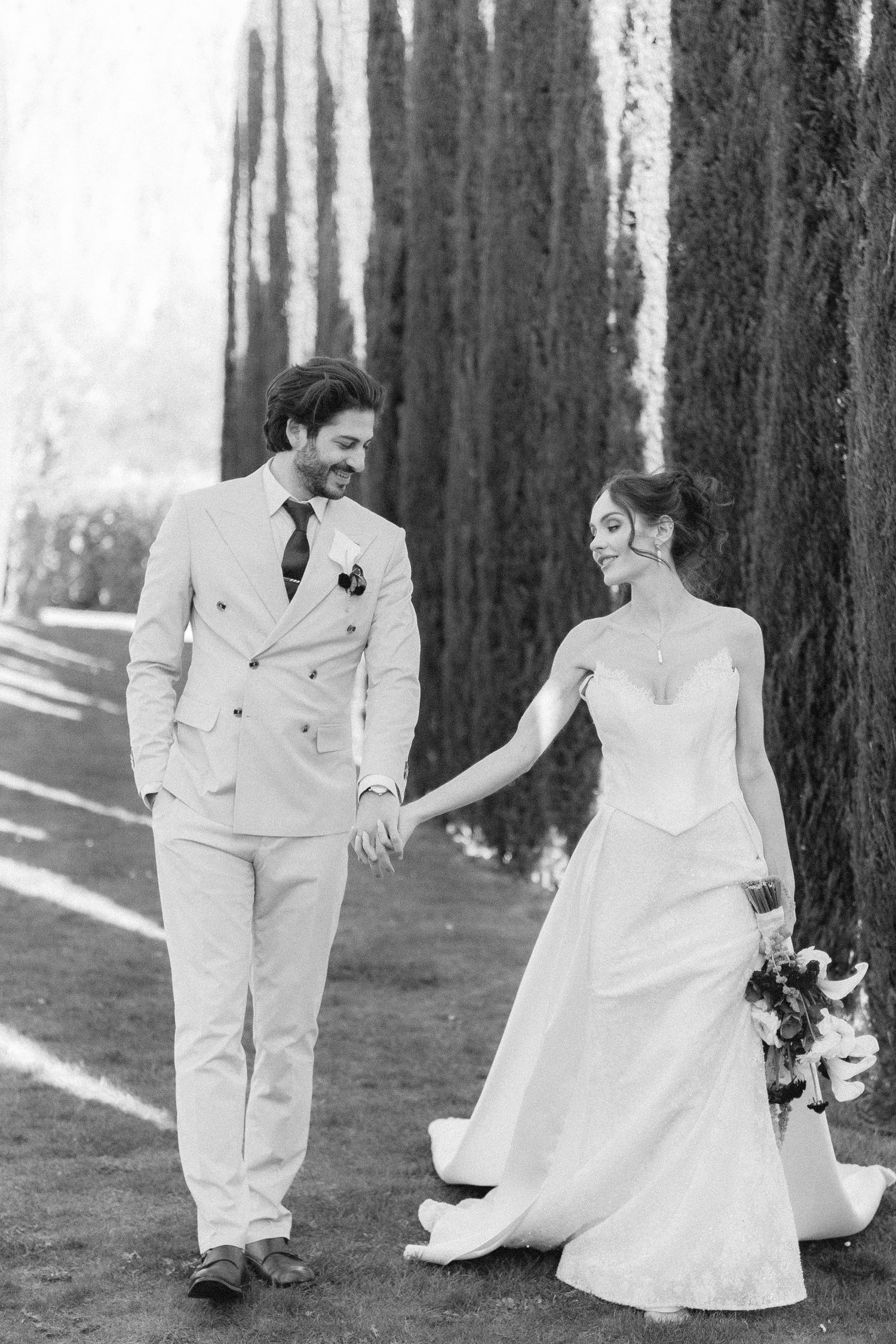Black and white photo of a bride and groom holding hands while walking among tall cypress trees on their wedding day.