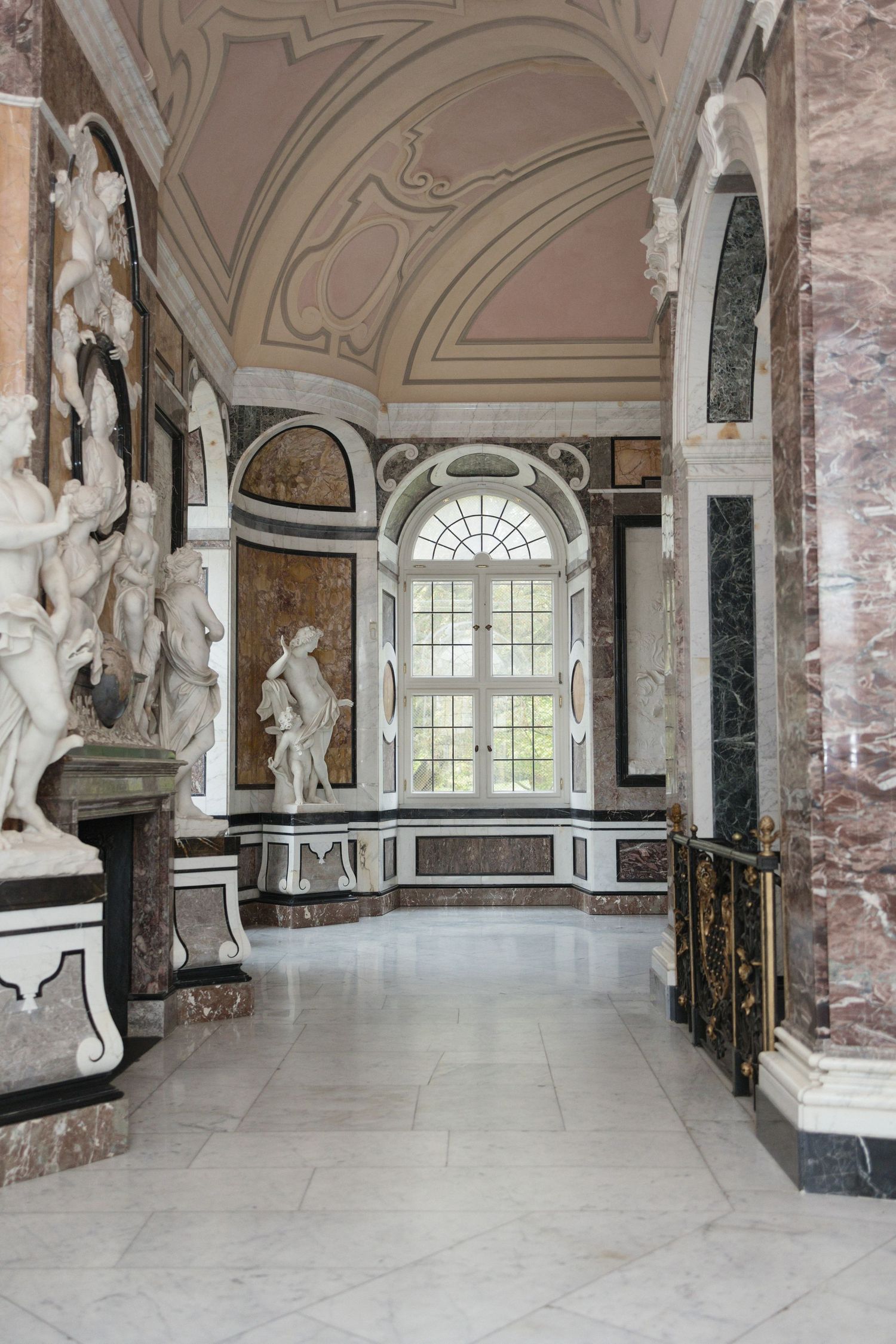 Ornate baroque palace interior with marble floors, white sculptures, decorative columns, and an arched window flooding the hall with light.