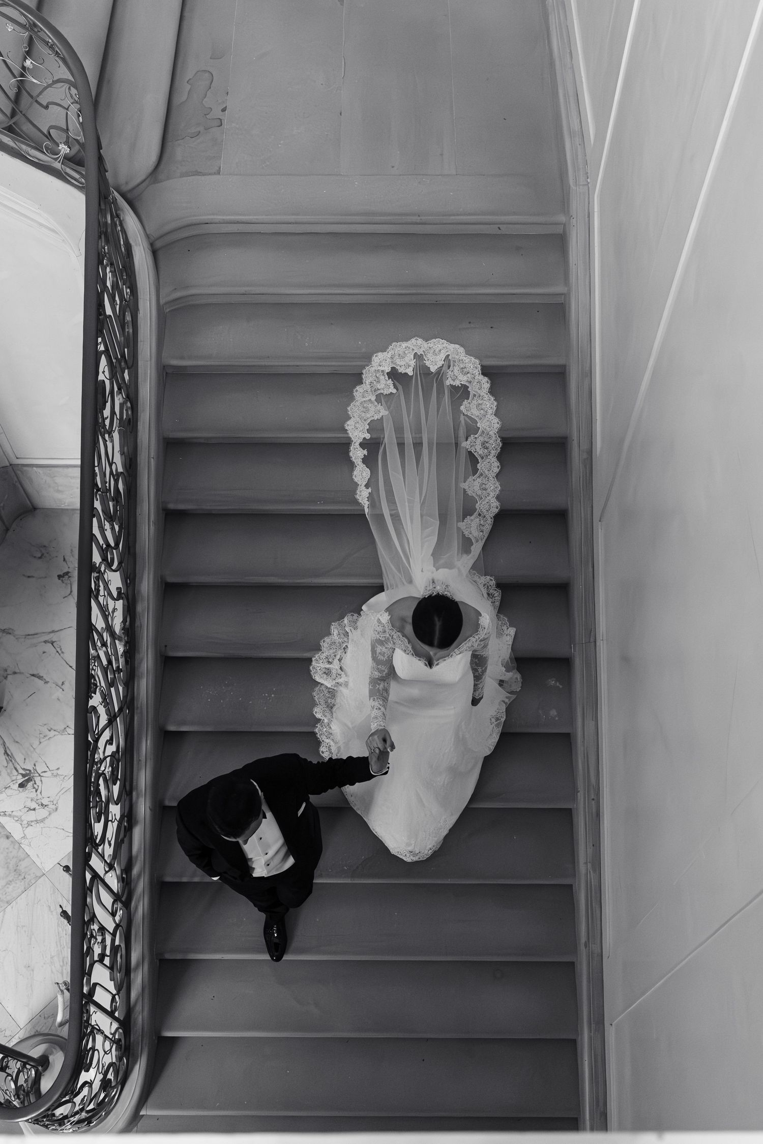 Black and white aerial view of bride and groom ascending grand staircase, bridal veil cascading elegantly.
