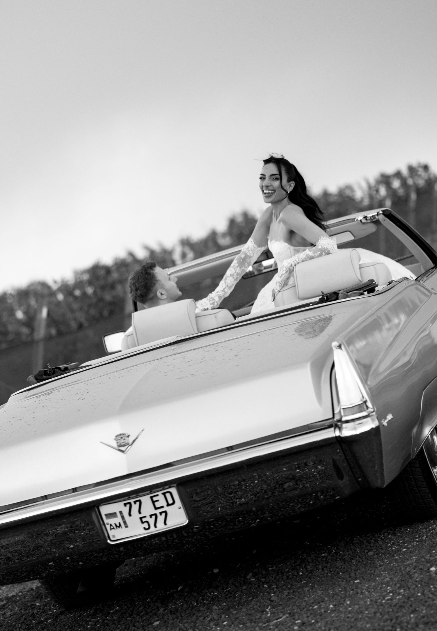 Black and white photo of a woman in a white dress posing in a classic convertible car with license plate 77 ED 577.