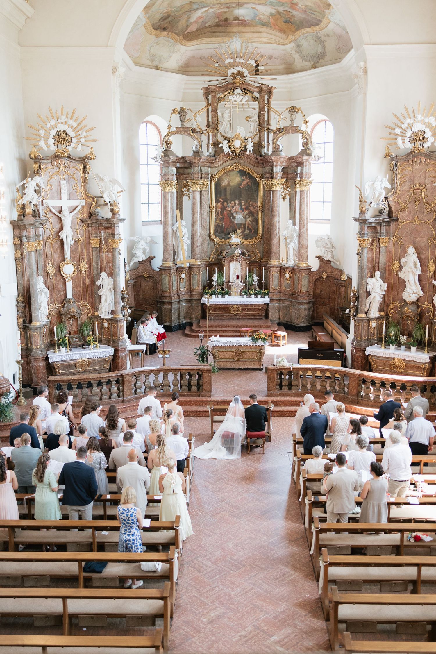 Elegant wedding ceremony in ornate Baroque church with guests seated in wooden pews as bride and groom stand at altar.