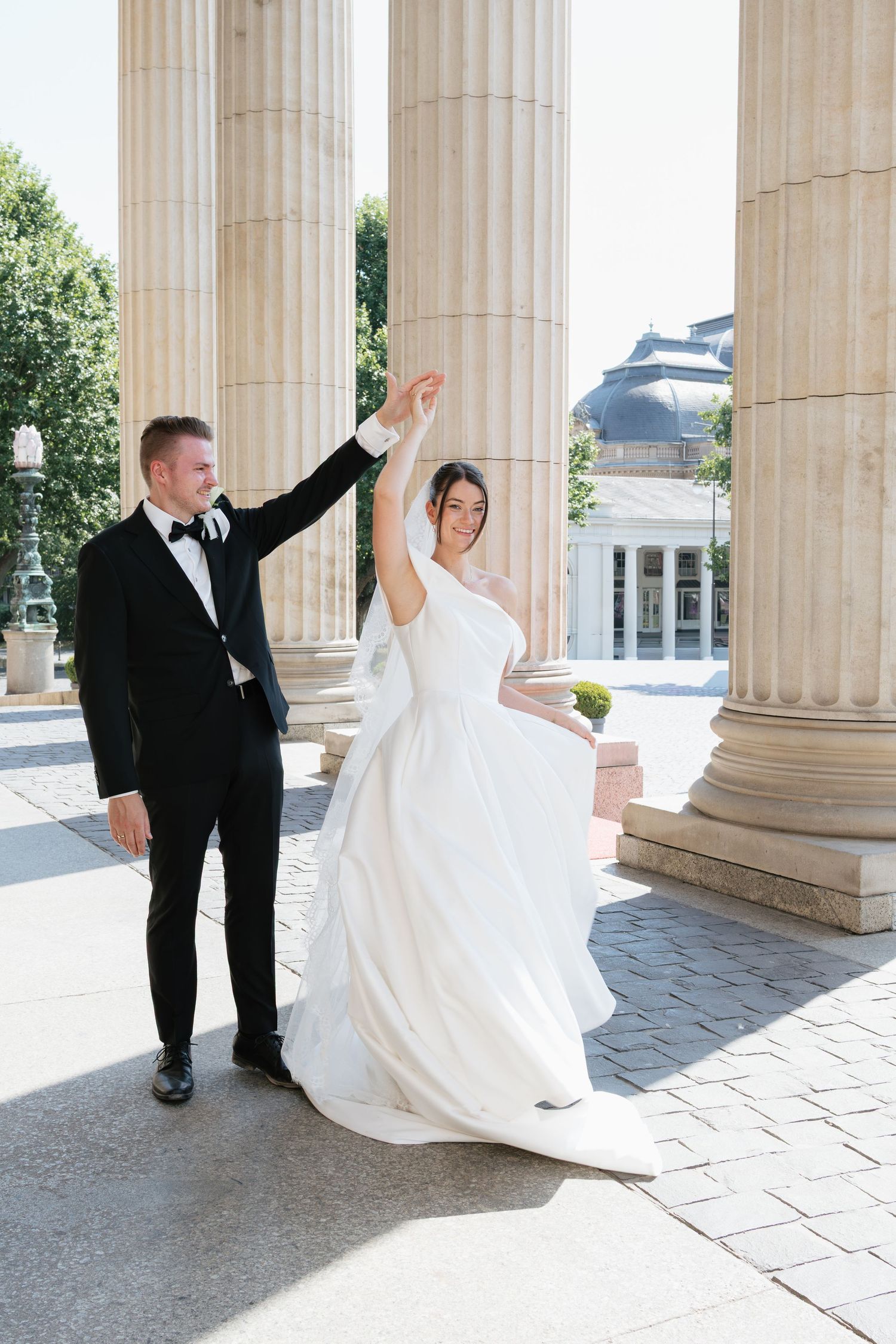 Bride in white gown twirls as groom in tuxedo holds her hand near grand stone columns at elegant wedding venue.
