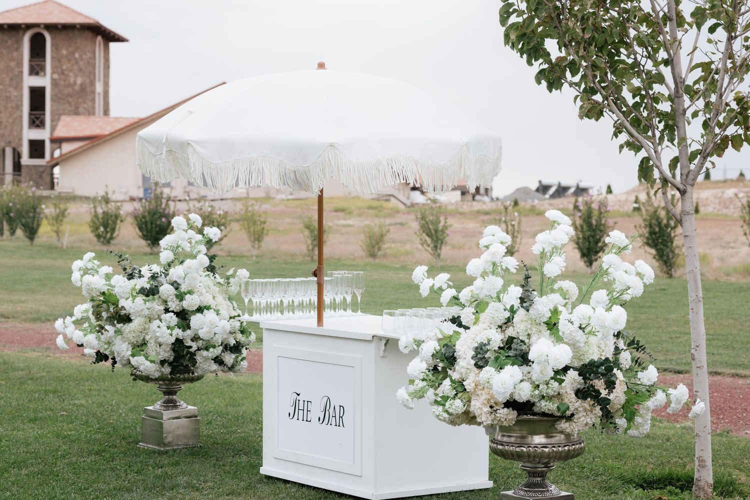 Elegant outdoor wedding setup with white floral arrangements and a white bar cart on a green lawn.