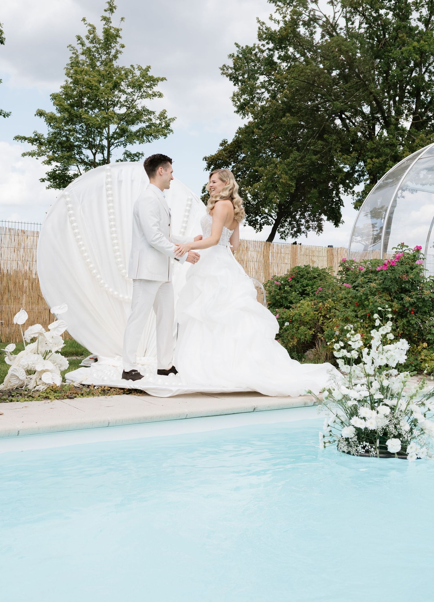 Bride and groom stand holding hands by a pool beside a large white shell backdrop with white floral arrangements.